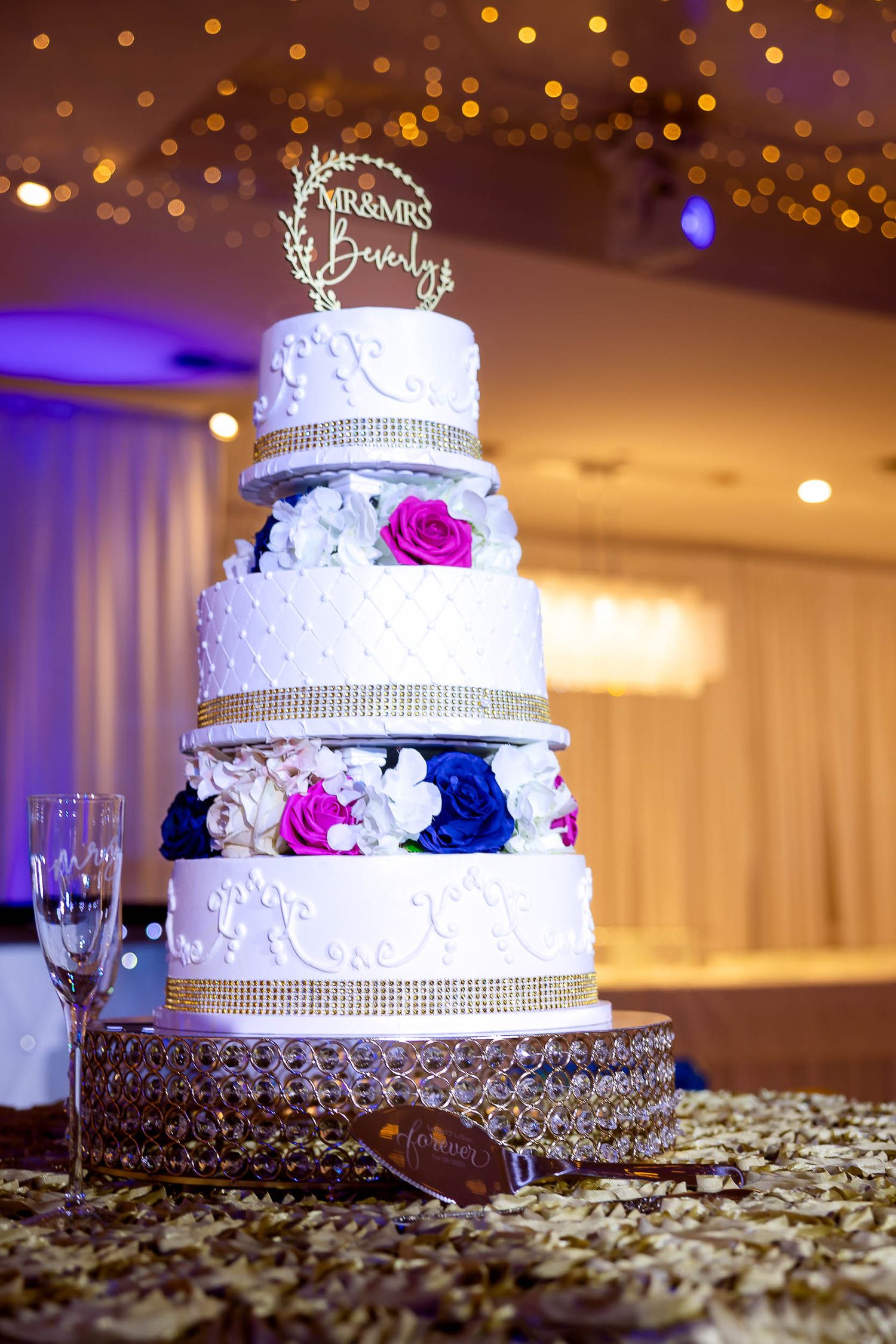 Elegant five-tier white wedding cake with crystal bands, pink and blue roses, and ornate silver cake topper on crystal stand.