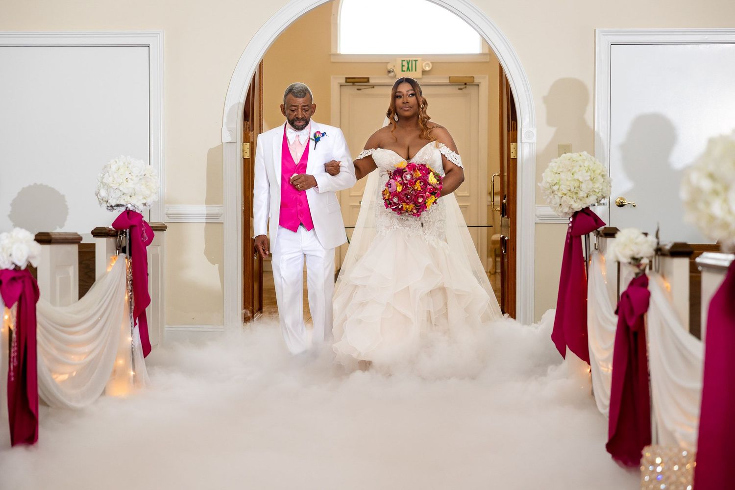 Wedding ceremony entrance decorated with white silk and burgundy ribbons as a couple stands under an elegant archway.