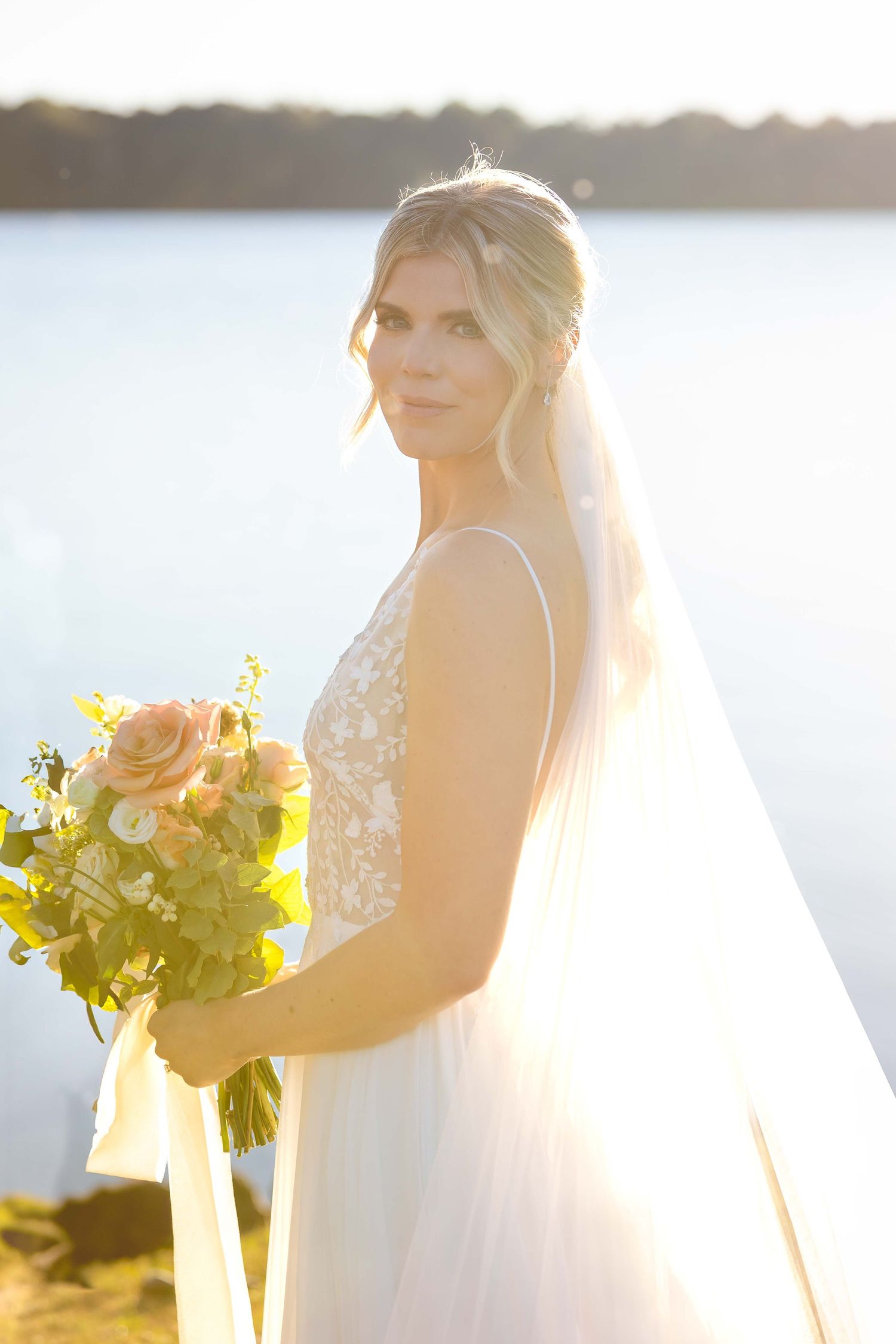 A bride in a white lace wedding dress holds a vibrant yellow and peach bouquet during golden hour by the water.