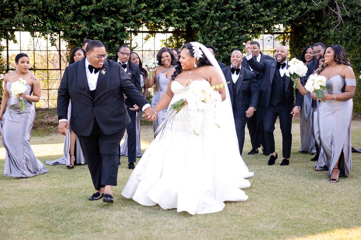 A wedding party in formal attire walks on a grassy lawn during an outdoor celebration.