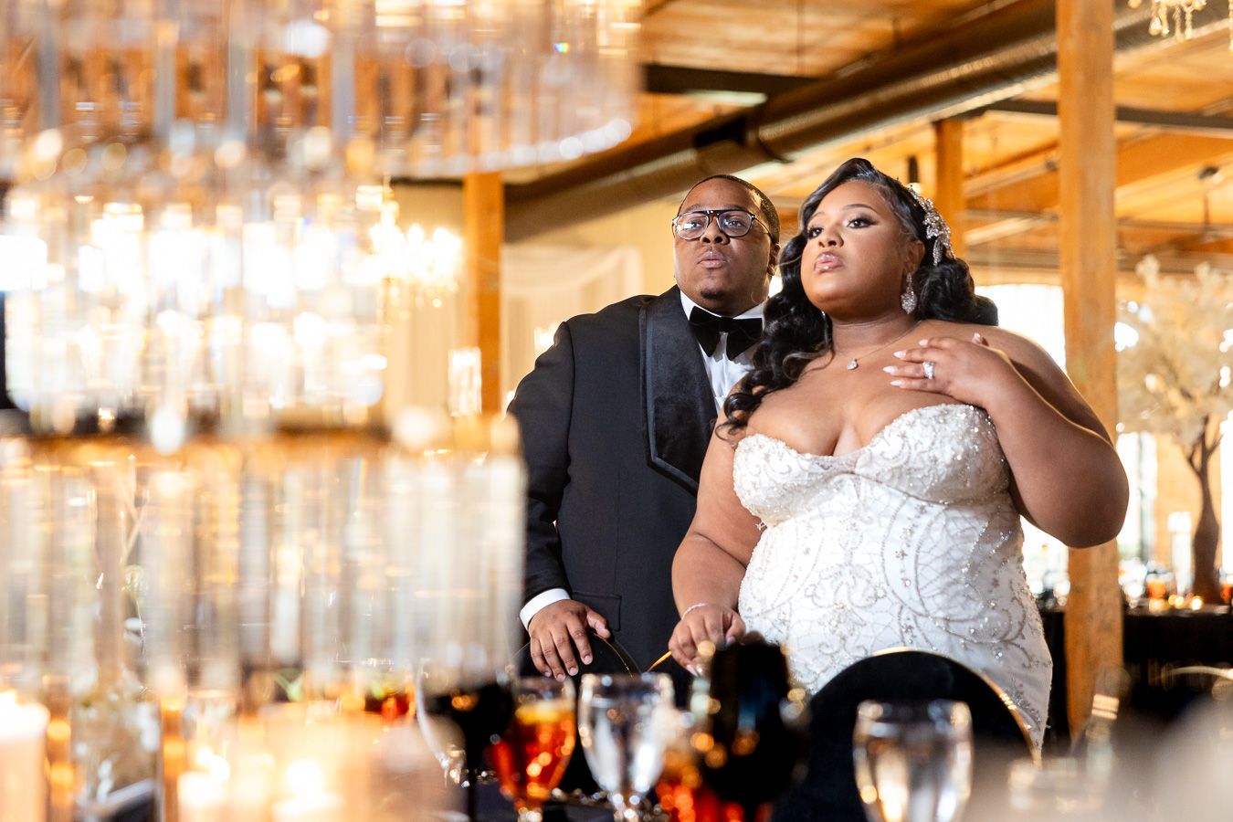 A couple in formal attire stands together at an elegant bar with crystal chandeliers hanging overhead.