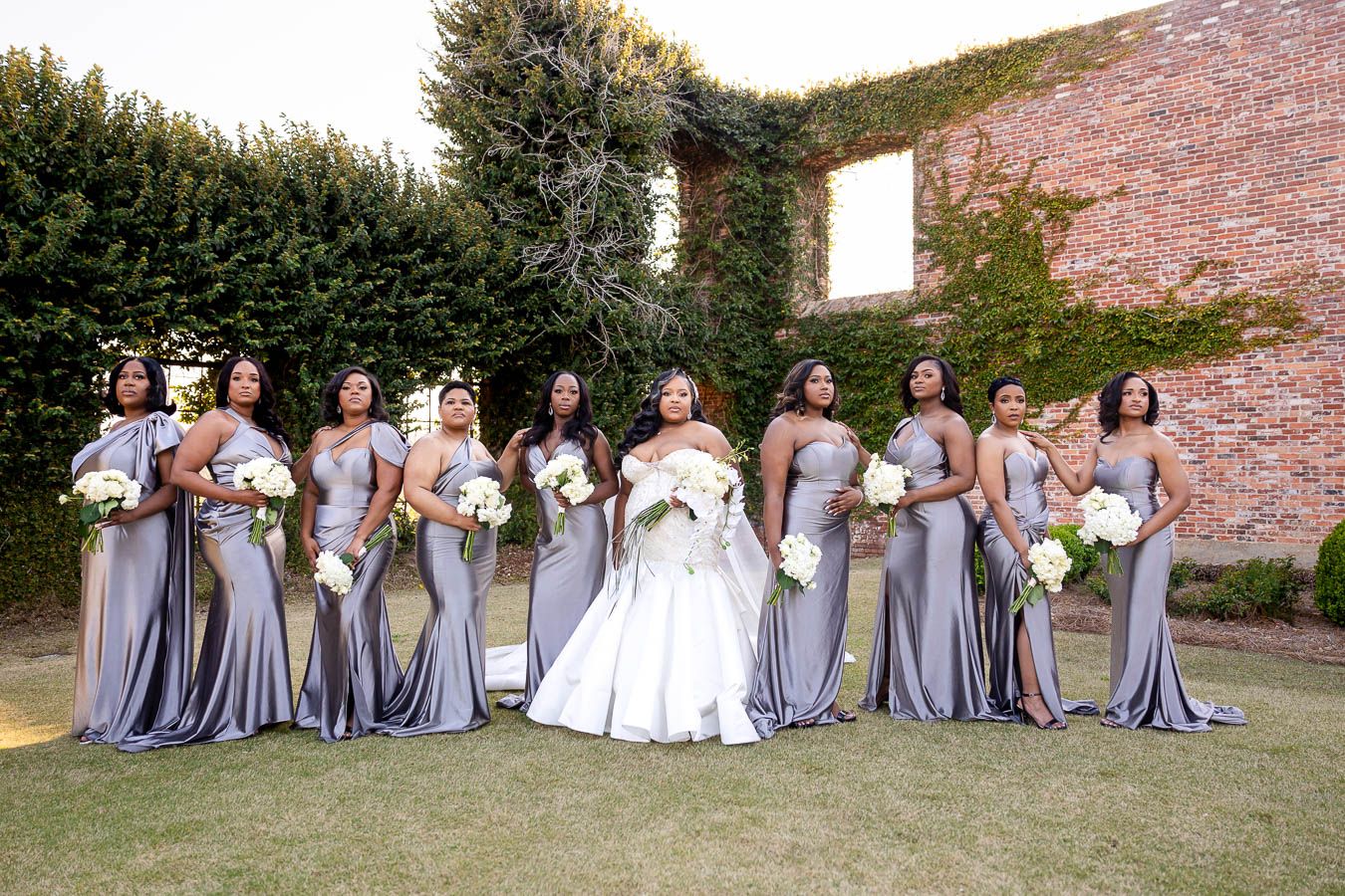 Wedding party wearing lavender bridesmaid dresses poses together against rustic brick wall with greenery.