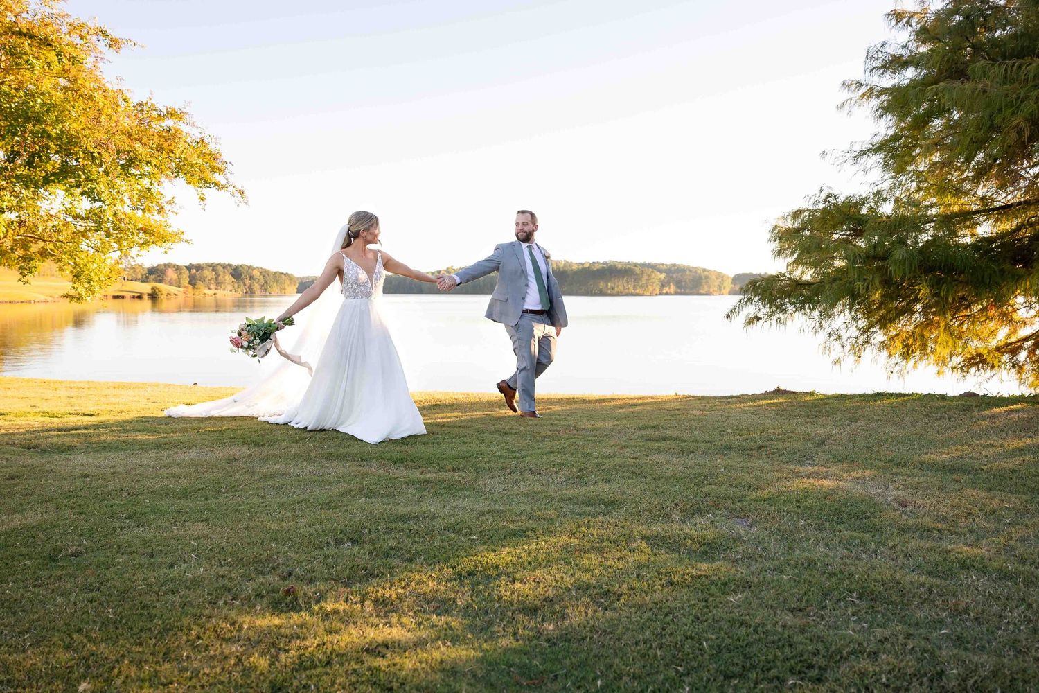 A couple dances joyfully on a grassy lawn at sunset beside a serene lake with golden autumn trees in the background.