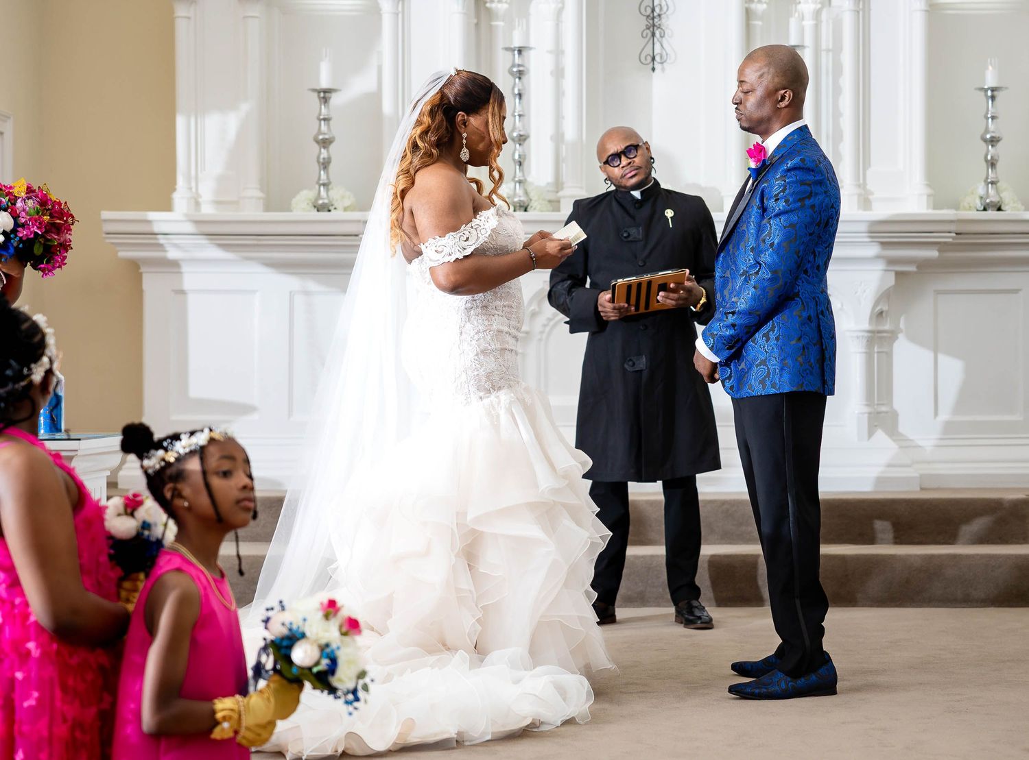 A wedding ceremony takes place in a bright church sanctuary with elegant white columns and floral decorations.