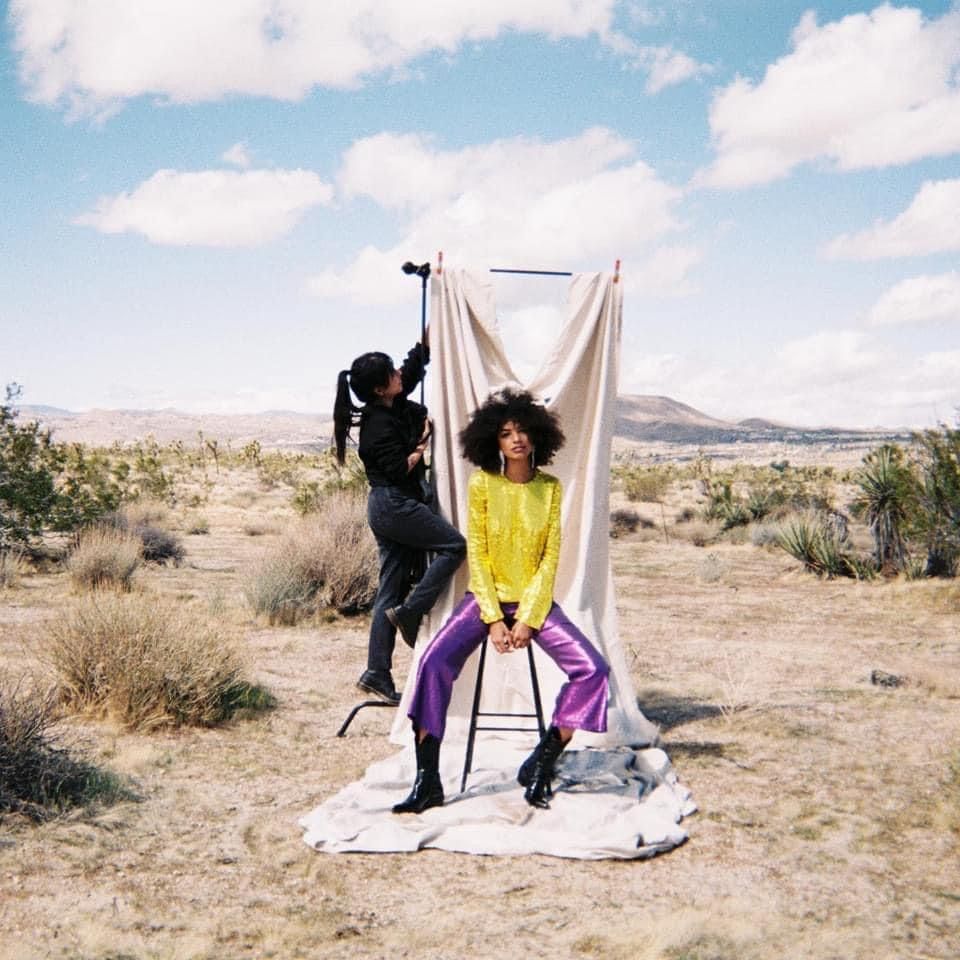 Behind-the-scenes fashion photoshoot in desert landscape with white backdrop and camera equipment.