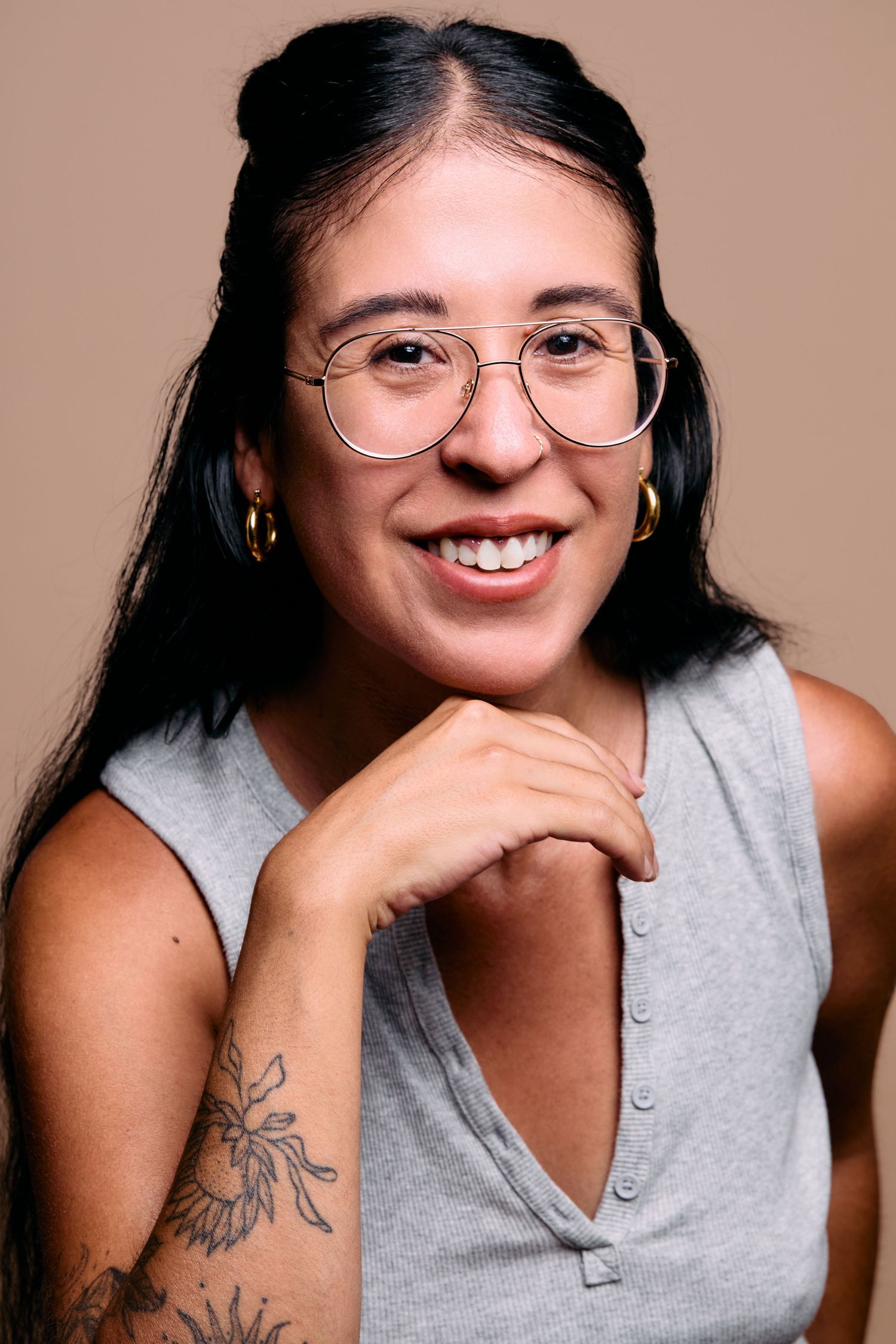 Person wearing round glasses and grey sleeveless top smiles warmly while displaying floral forearm tattoo against neutral background.