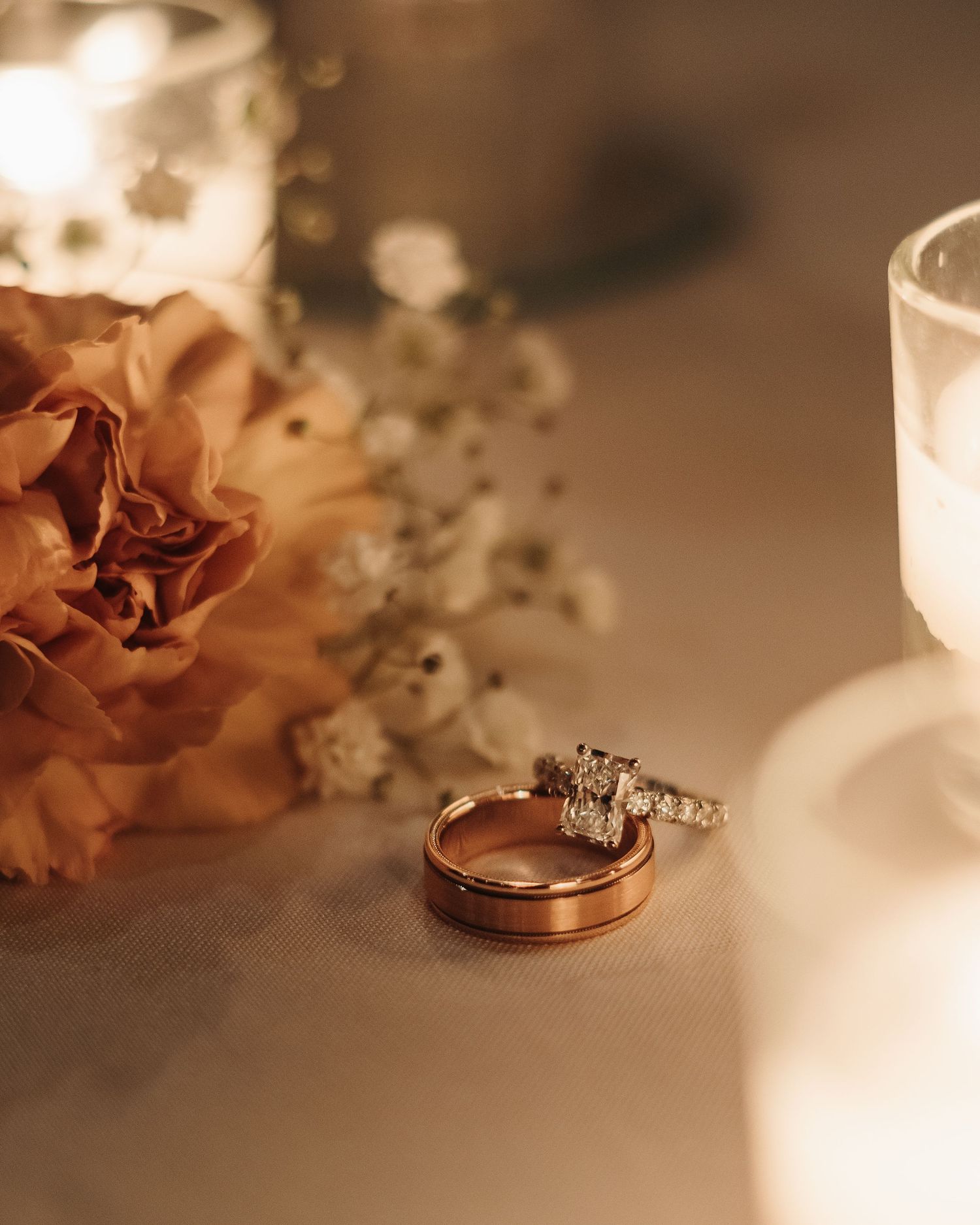 A close-up sequence showing gold wedding bands and delicate flowers arranged on a soft white surface in warm lighting.
