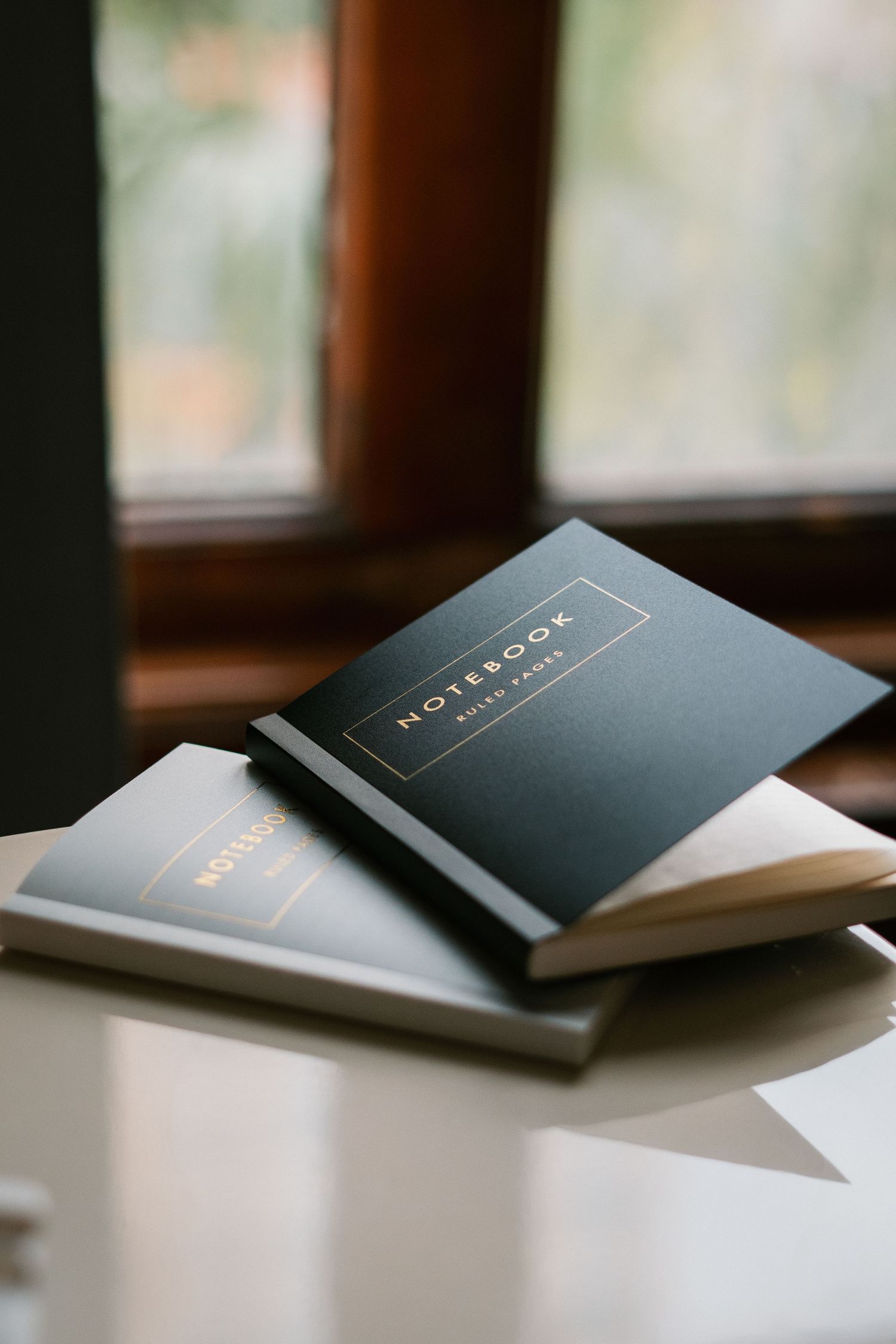 A series of black minimalist notebooks with gold lettering shown in progressive angles on a white surface near a window.