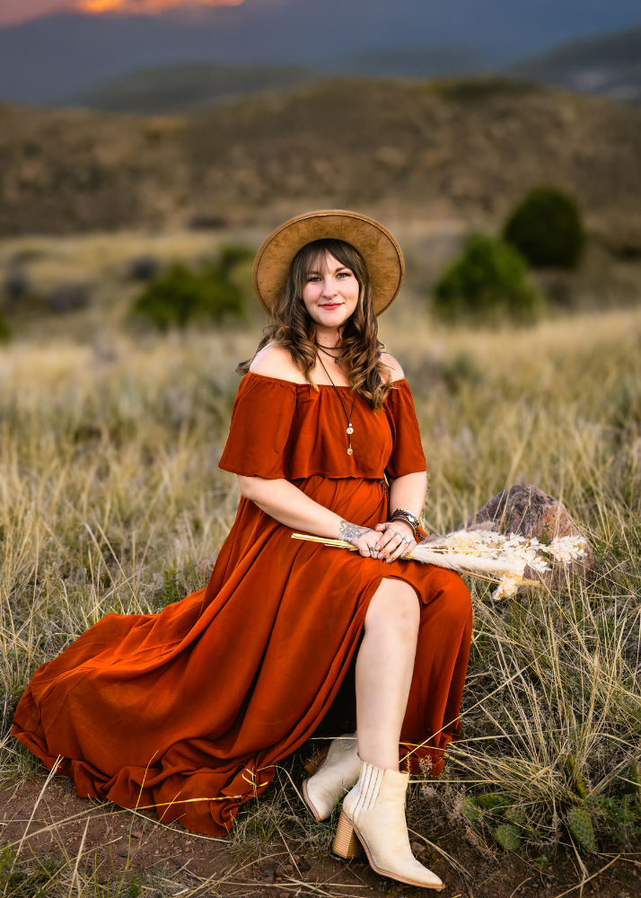 A maternity photographer in a rust-colored off-shoulder dress and felt hat poses on a log during sunset in a Colorado landscape.