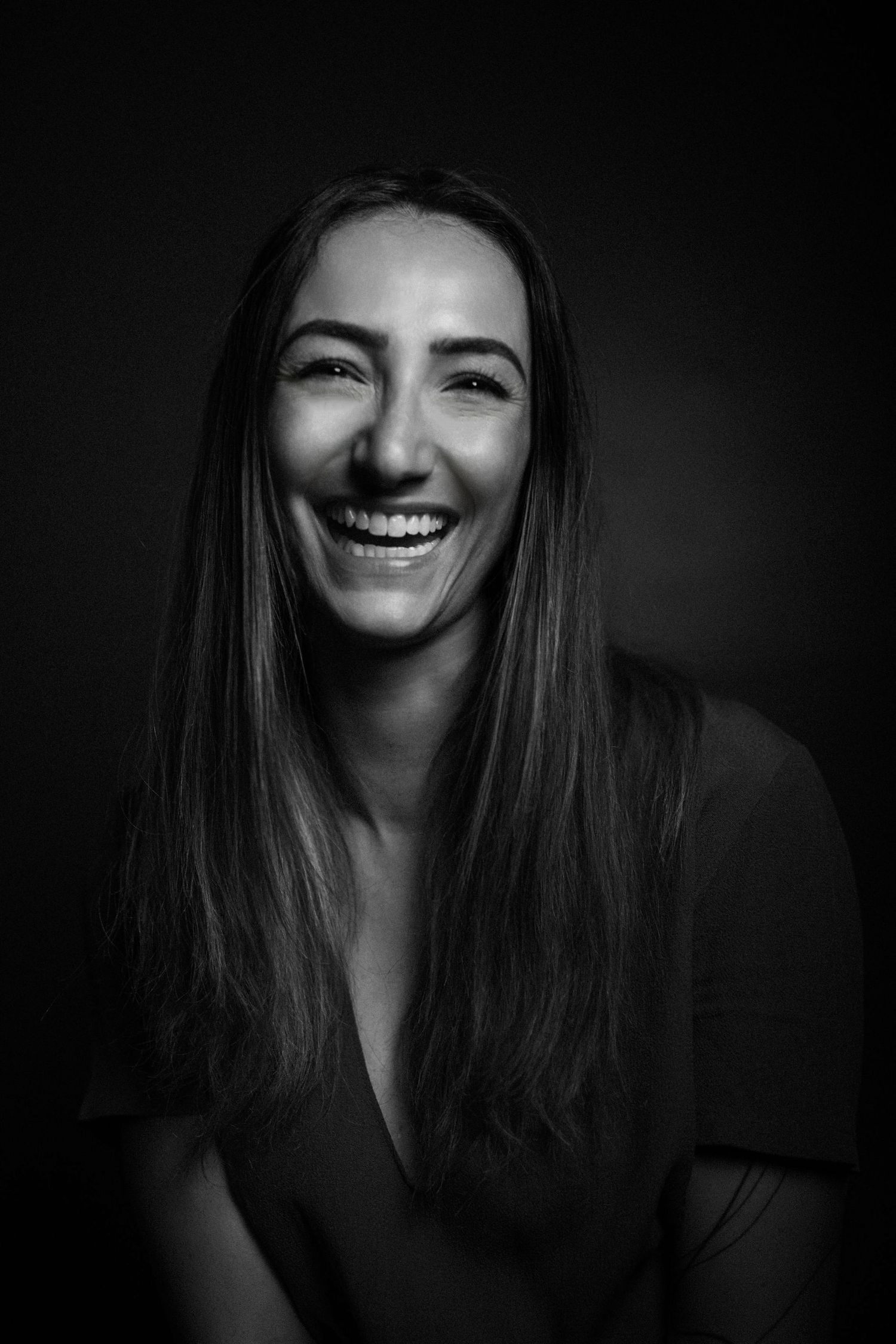 Black and white portrait photograph showing person with long dark hair smiling brightly against dark background.