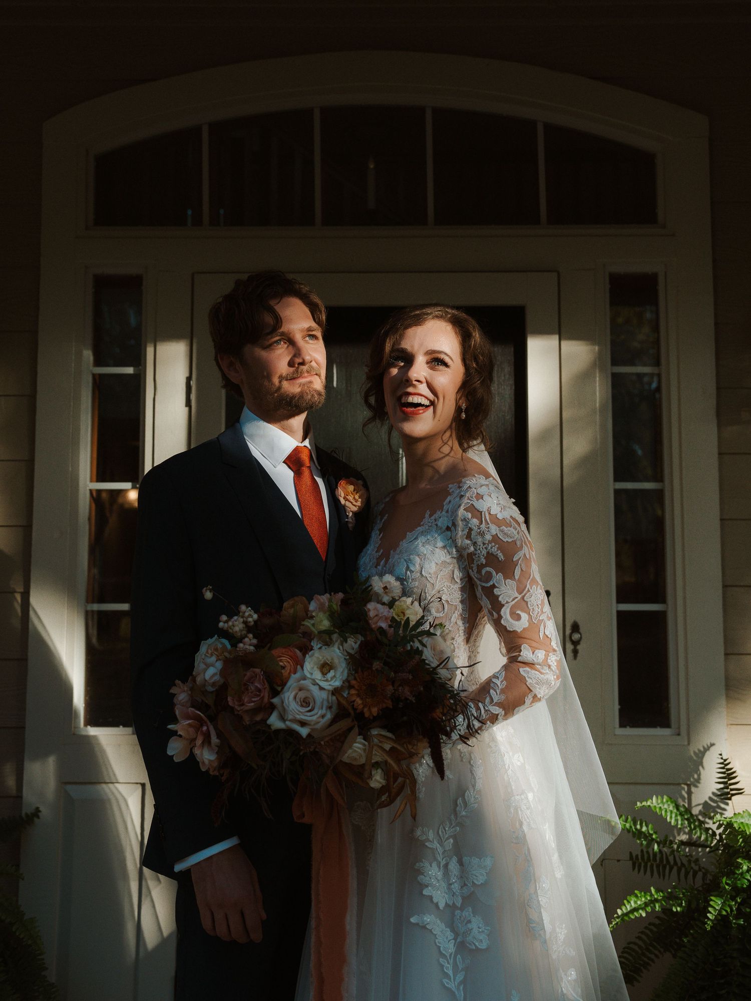 Sunlit wedding portrait in front of white doorway with dramatic autumn bouquet.