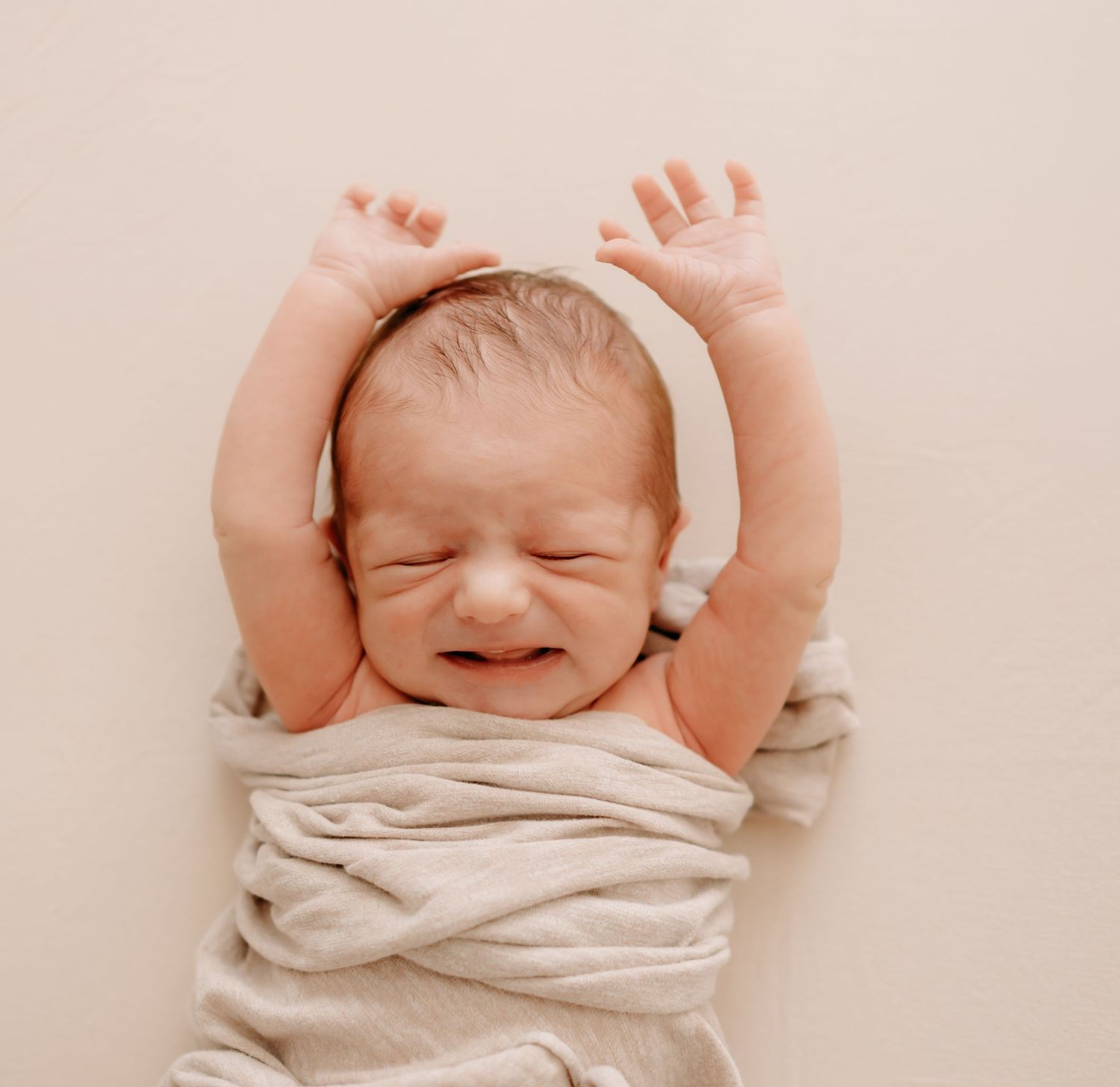 A newborn baby wrapped in beige cloth stretches arms up while making a joyful expression during professional newborn photos at home.