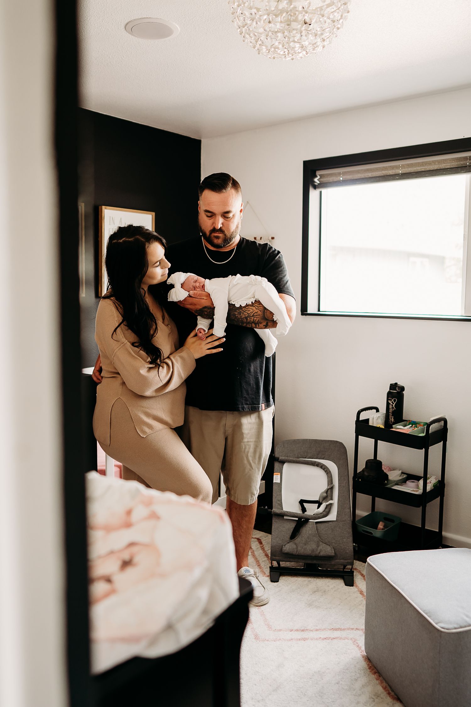 A couple stands in a bright nursery room holding their newborn baby while bathed in natural light from a window during in home photos.