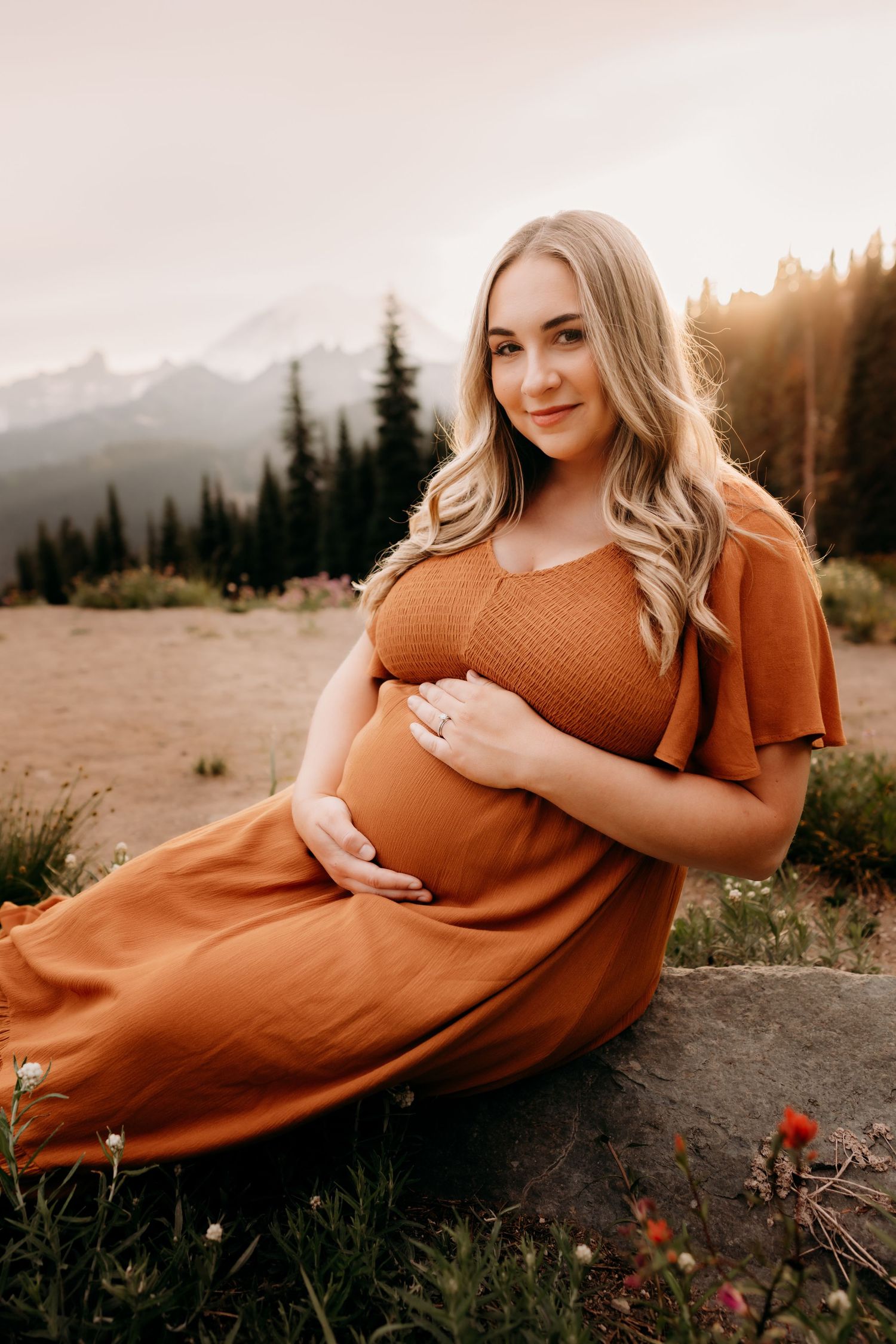 Pregnant woman sits on a rock surrounded with wildflowers with tall trees and Mt. Rainier in the background.