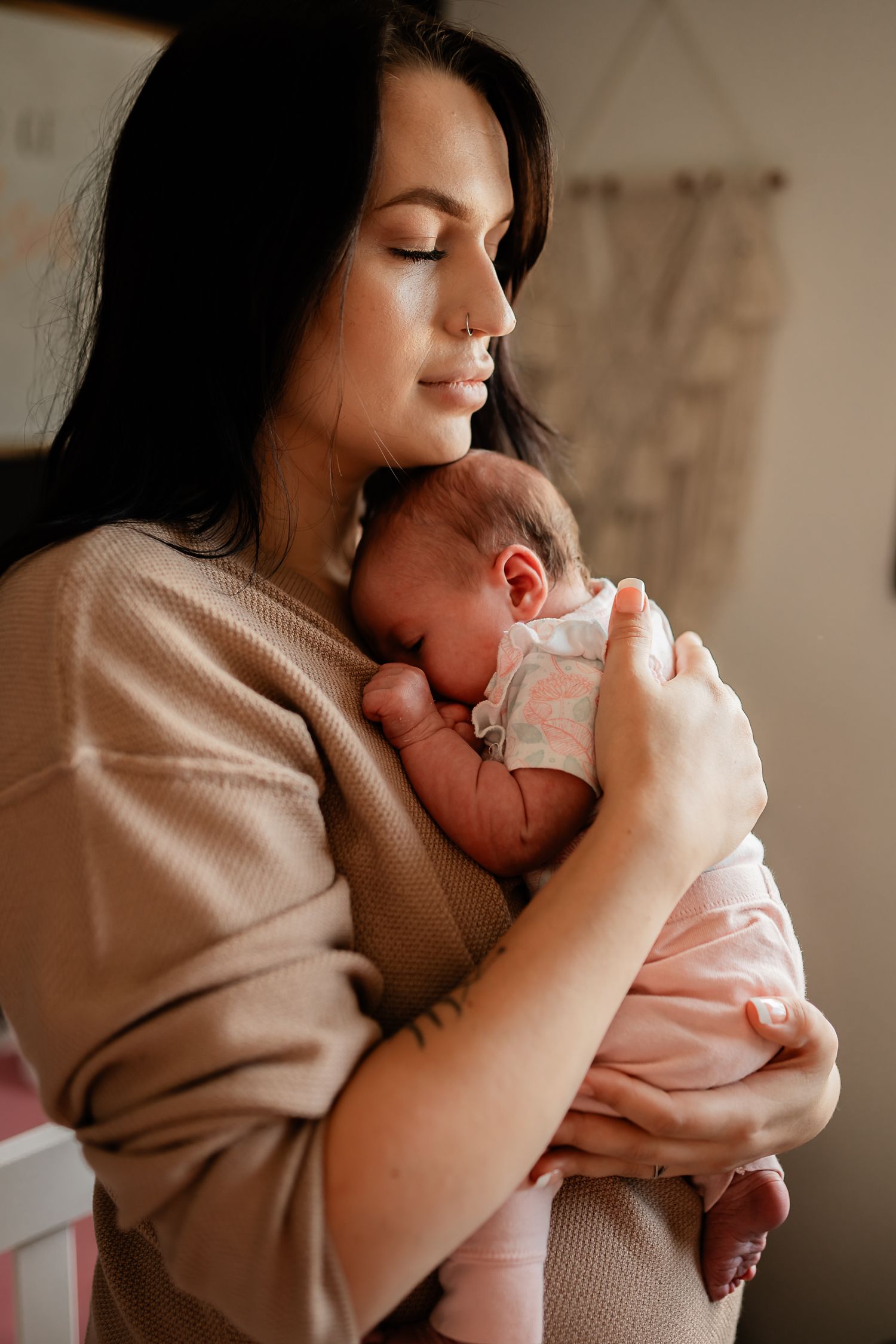 A tender moment shows a new mom in a beige sweater holding and gazing lovingly at their sleeping newborn in pink clothing.