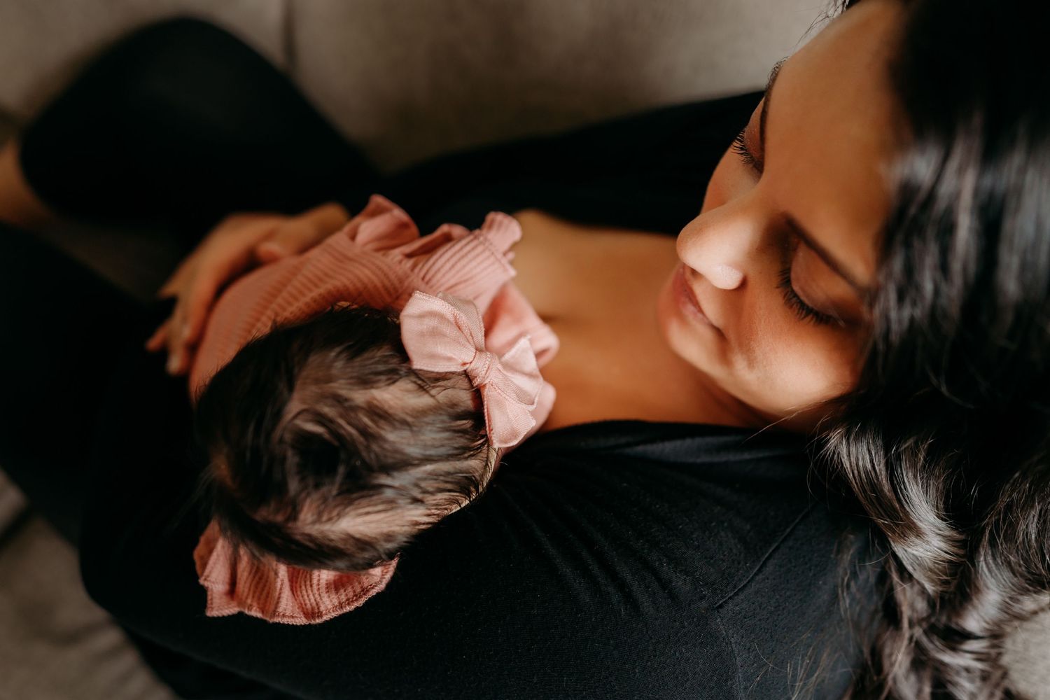A tender moment shows a mother in black clothing holding a sleeping baby wearing a pink bow headband.