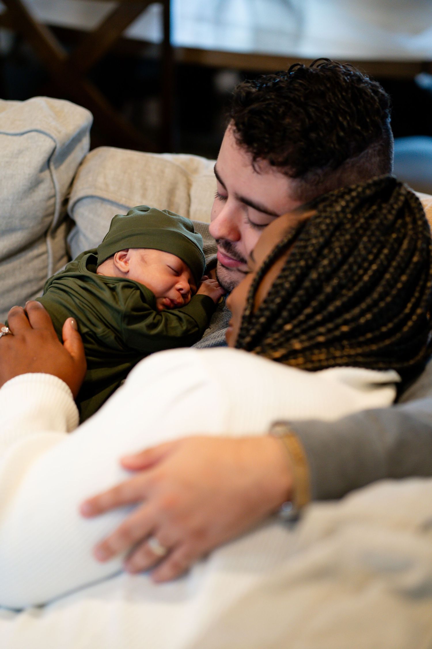 A newborn baby in green sleepwear rests peacefully on an dad's chest while relaxing on a couch during in home newborn photo session.