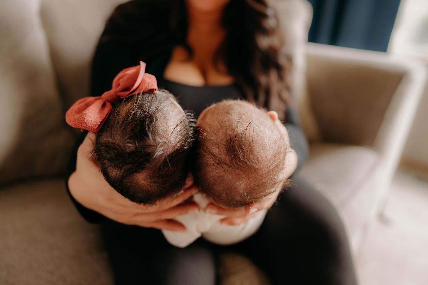 Two newborn babies sleeping while being cradled in arms on a beige couch during in home newborn photo session in Puyallup.