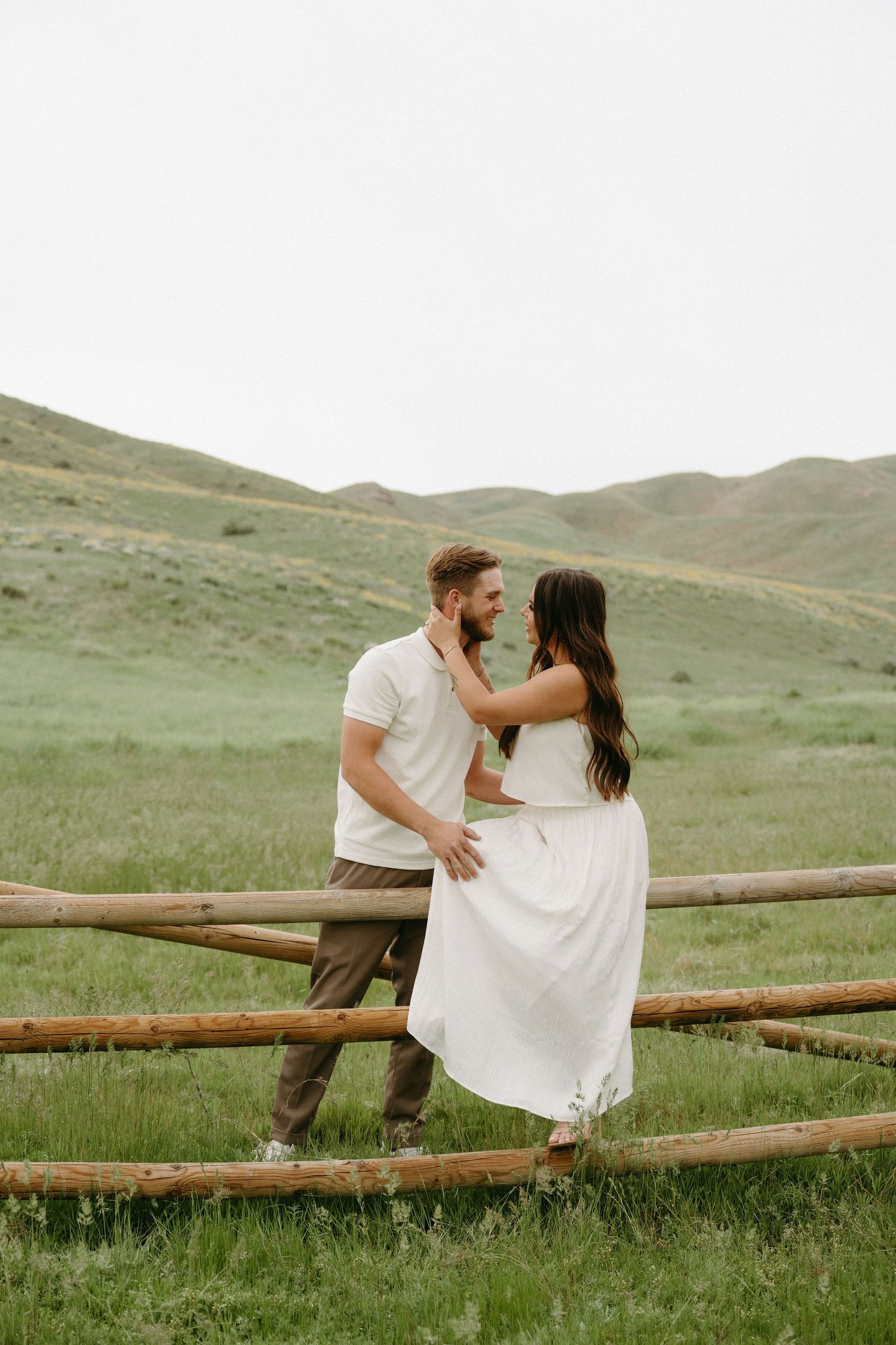 A couple shares a romantic moment while sitting on a wooden fence in a green mountainous meadow.