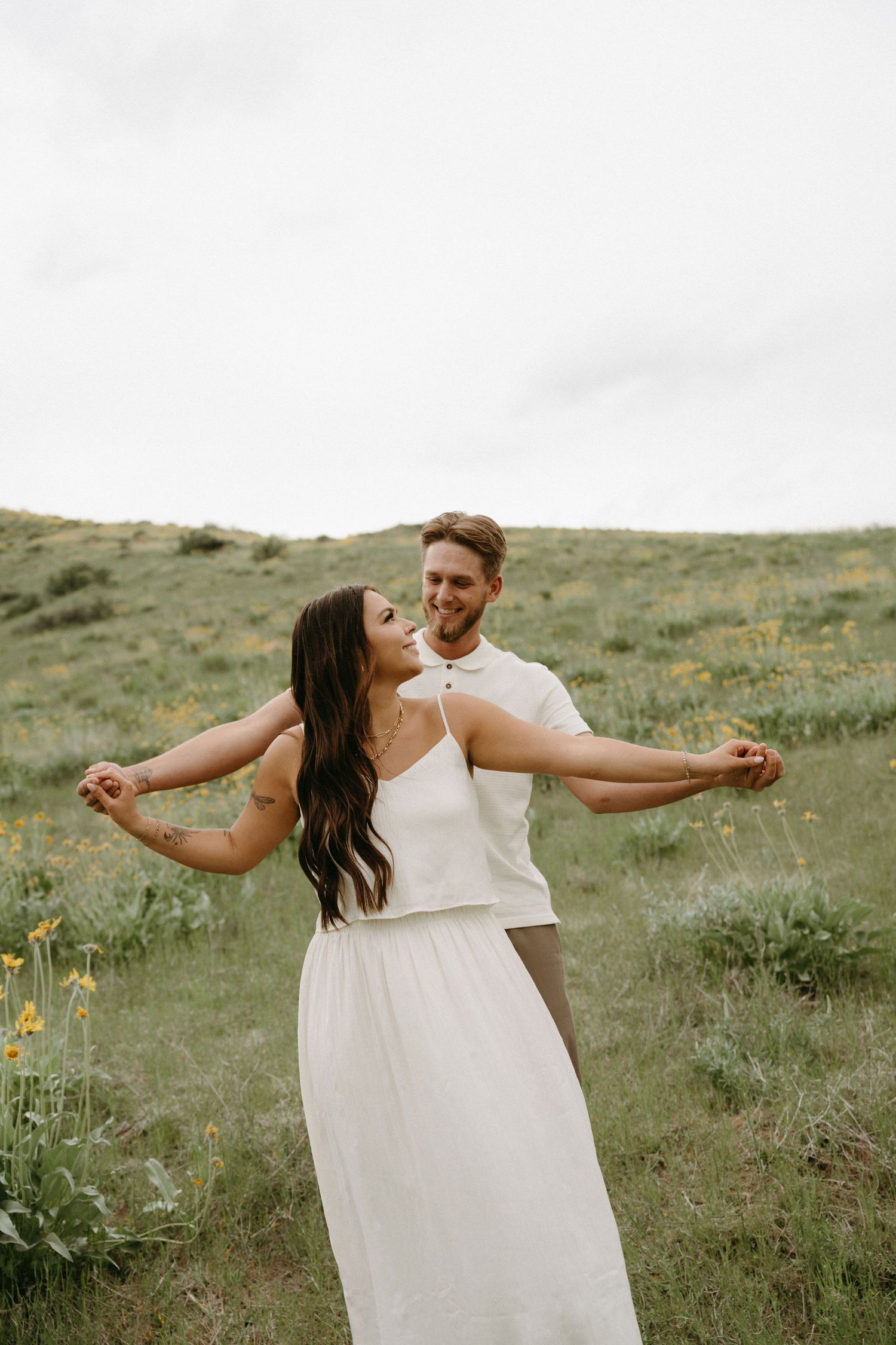 A couple dances playfully in a meadow with wildflowers and hills in the background.