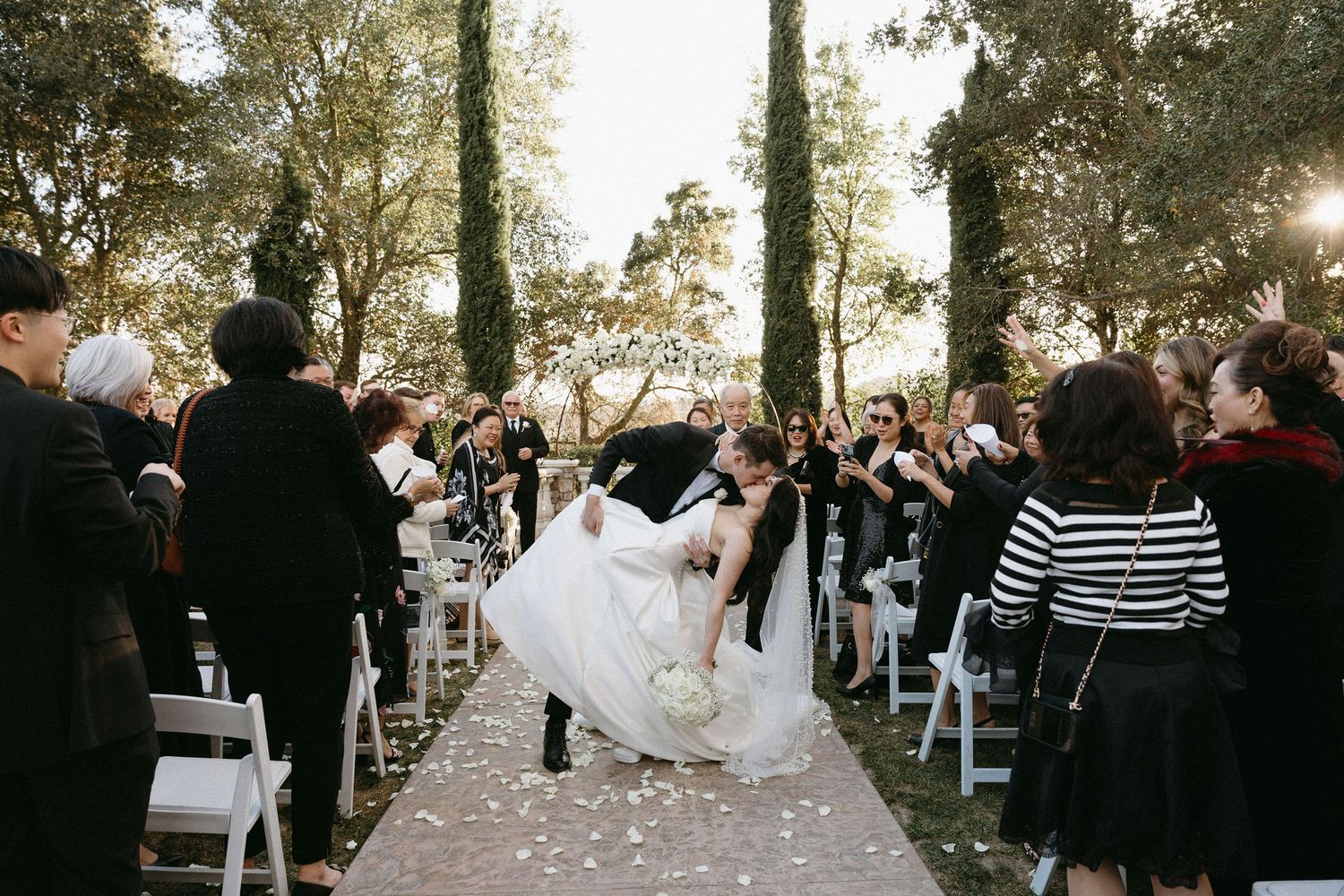 A joyful couple shares their first kiss at an outdoor garden wedding ceremony while guests look on and celebrate.
