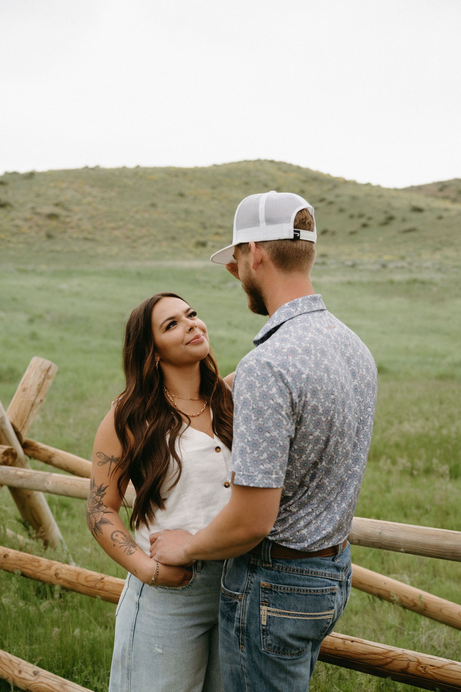 A couple shares a tender moment while leaning against a wooden fence in a rural mountain setting.