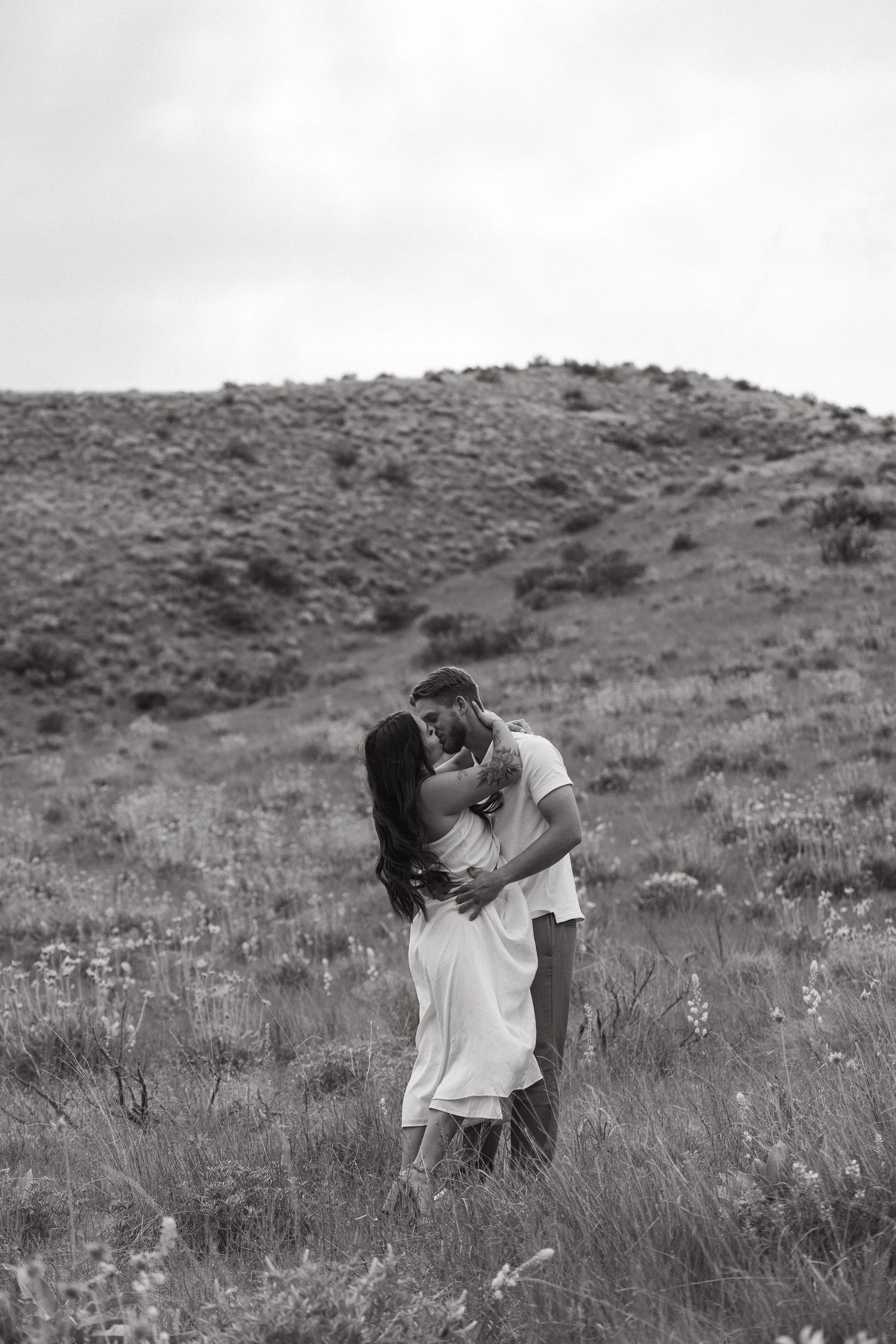 A romantic couple embraces in a black and white photo on a hillside covered in wild grass and flowers.