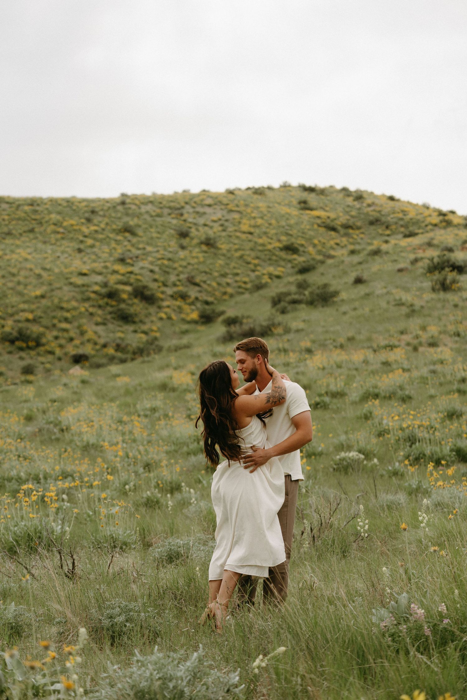 A couple shares a tender moment together in a green meadow filled with wildflowers on a hillside.