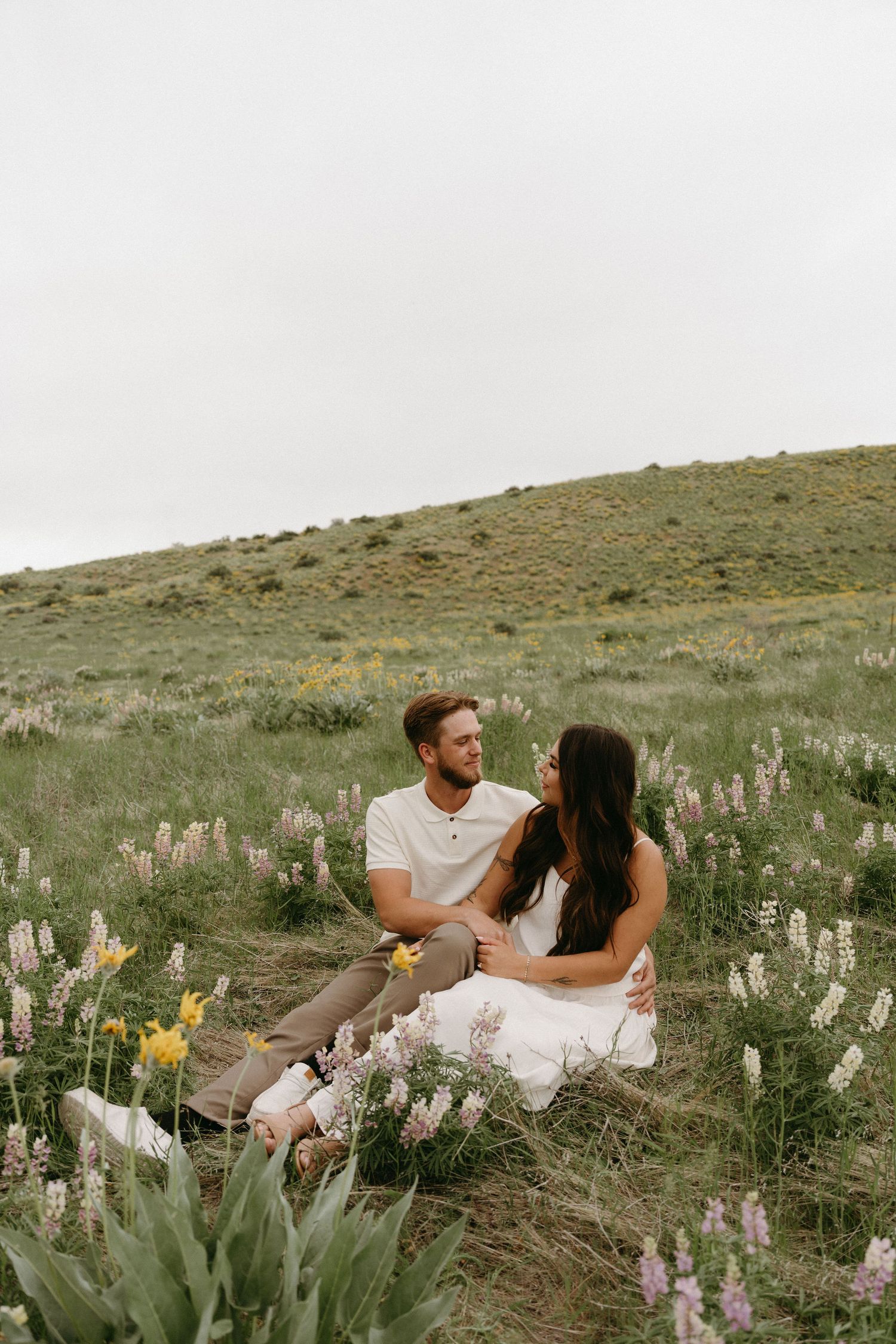 A couple sits together in a field of wildflowers on a grassy hillside during a romantic outdoor photoshoot.