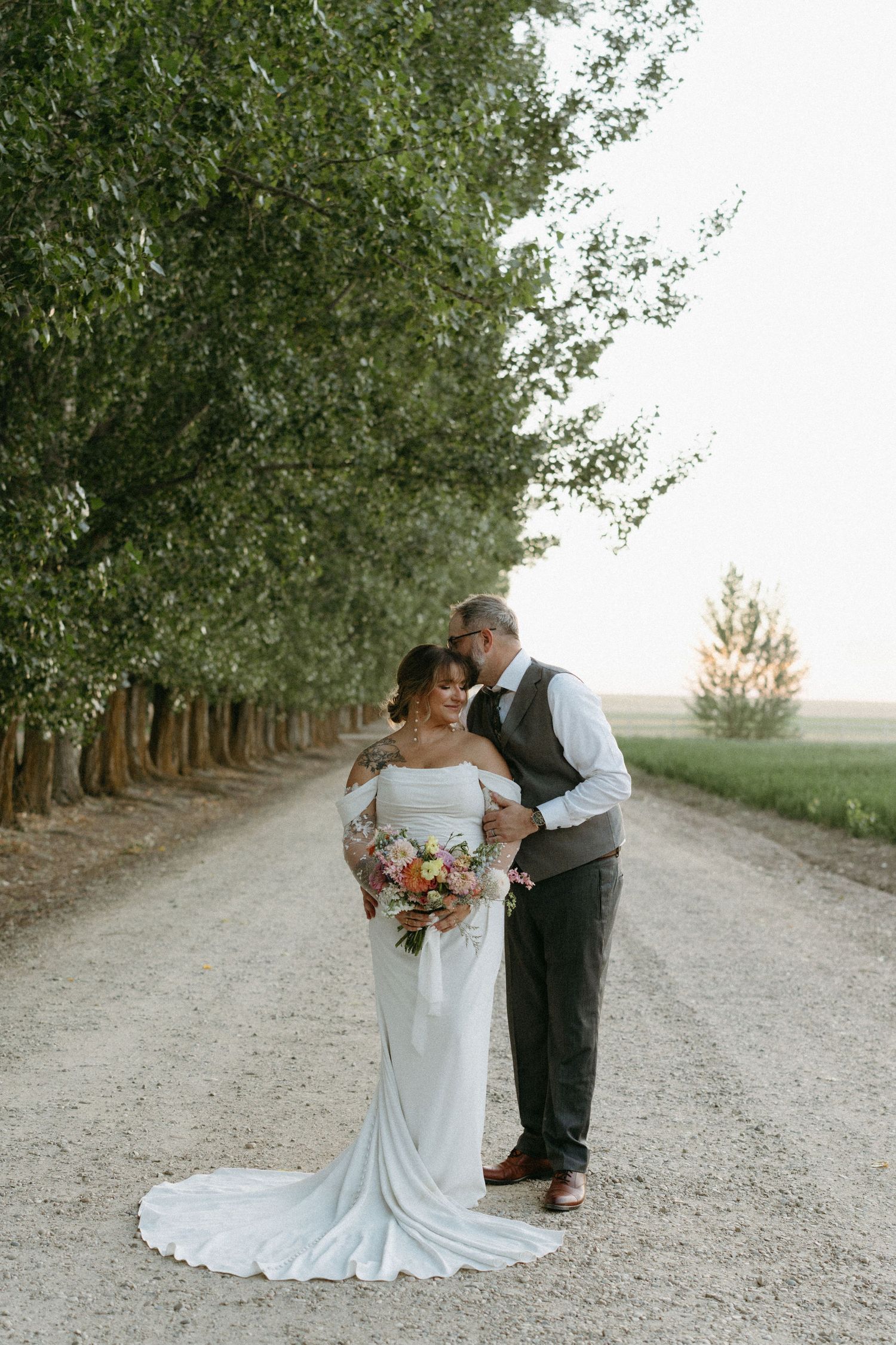 A couple shares a romantic moment on a tree-lined dirt road during their wedding photo session at sunset.