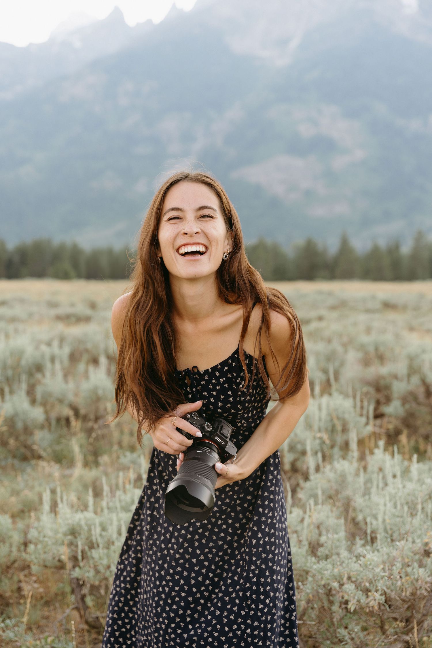 A joyful photographer wearing a polka dot dress laughs while holding a camera in a mountain meadow at sunset.
