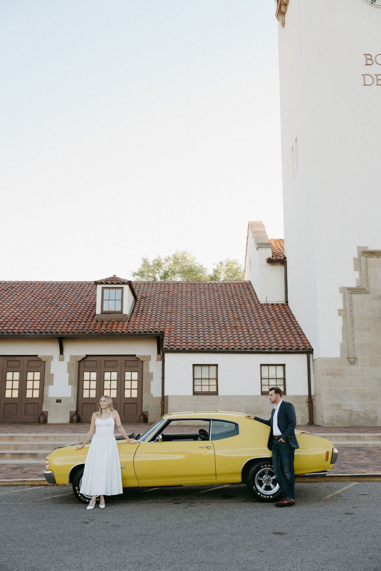 A couple stands next to a vintage yellow muscle car parked in front of a white building with terracotta roof tiles.