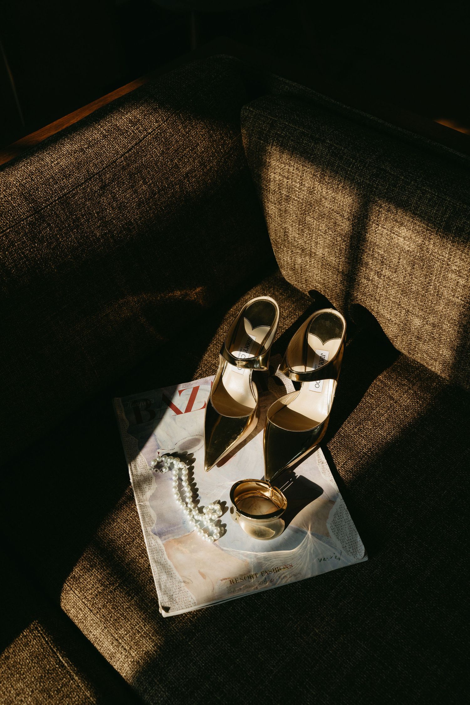 Wedding accessories including elegant heels and jewelry arranged on a tan couch in dramatic lighting.