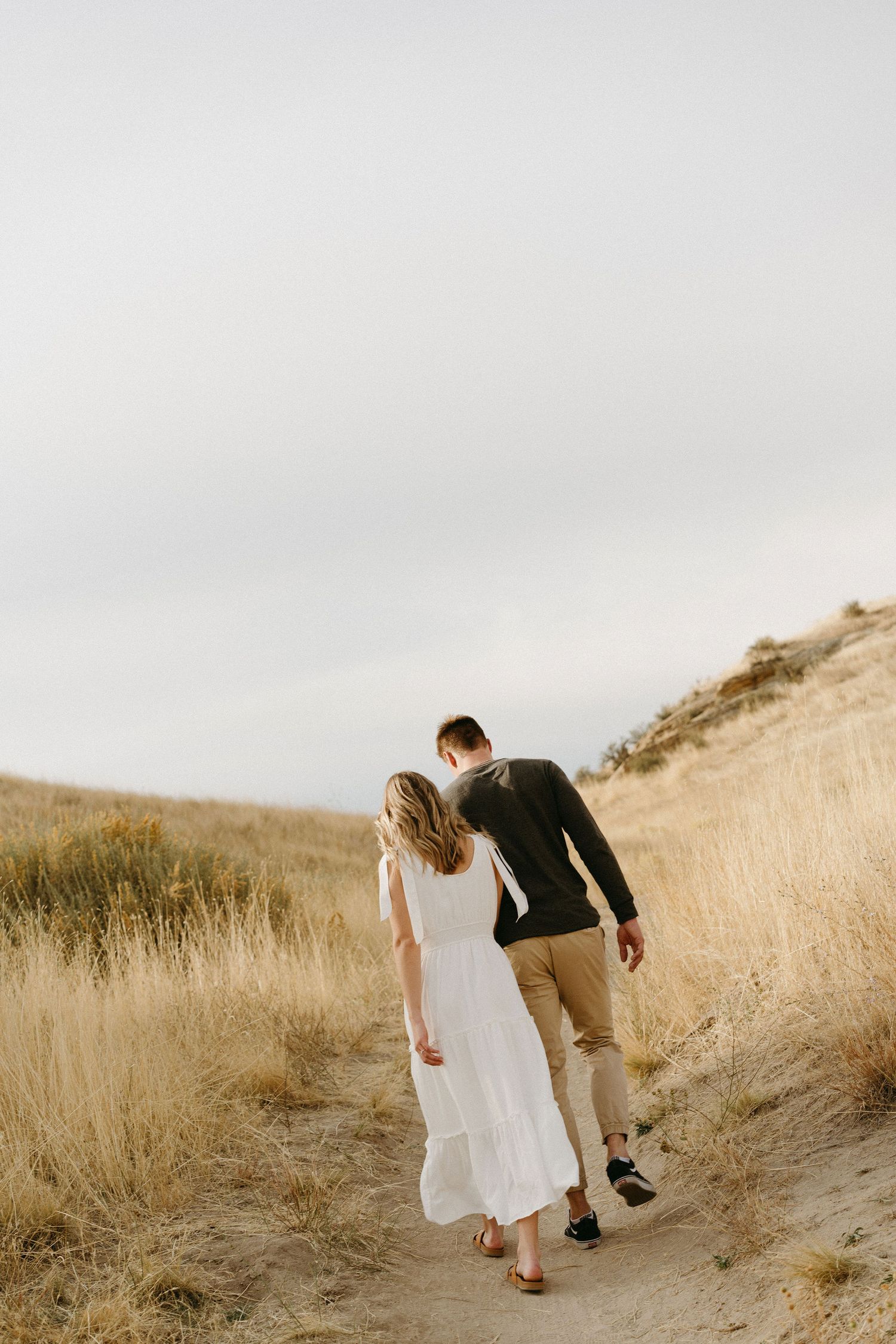 A couple walks together on a golden hillside path during sunset, with her flowing white dress swaying in the breeze.
