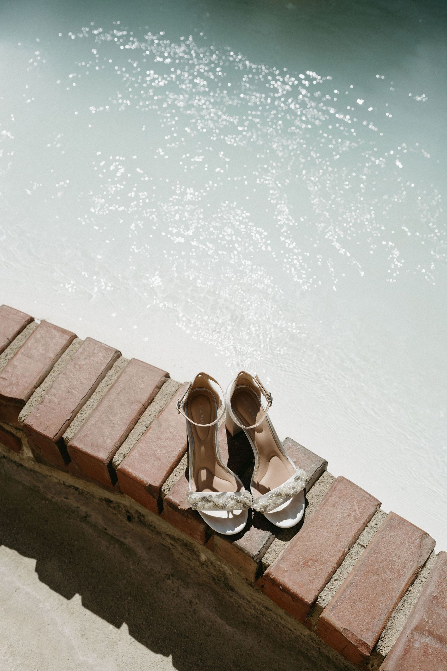 A pair of elegant silver bridal sandals sit on a brick ledge beside turquoise water at a poolside.