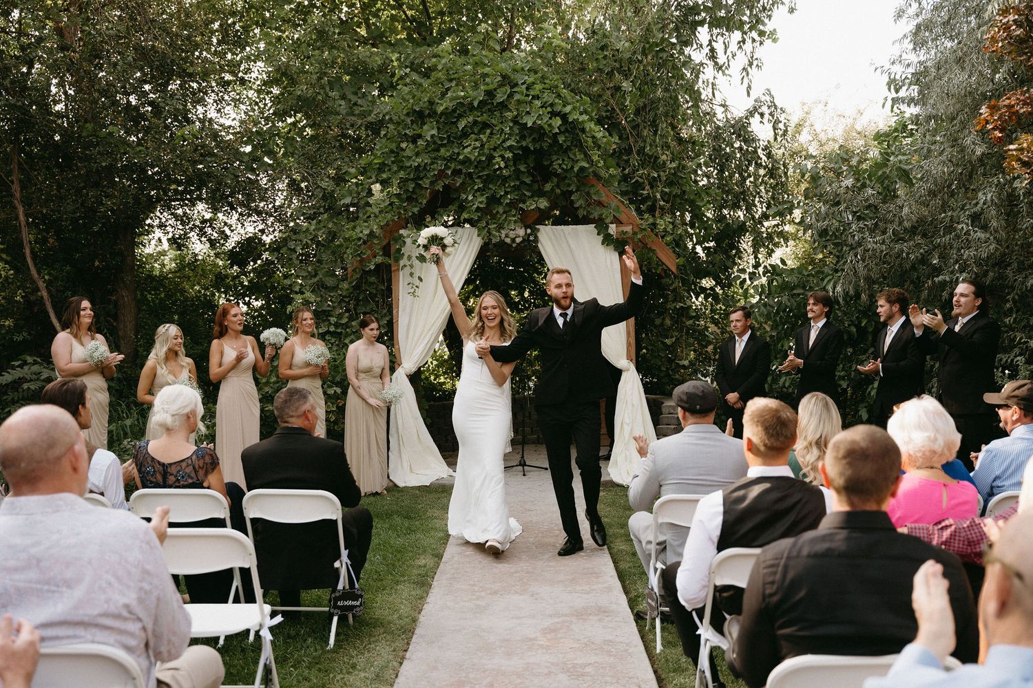An outdoor wedding ceremony takes place under a floral arch on a stone path surrounded by trees and seated guests.