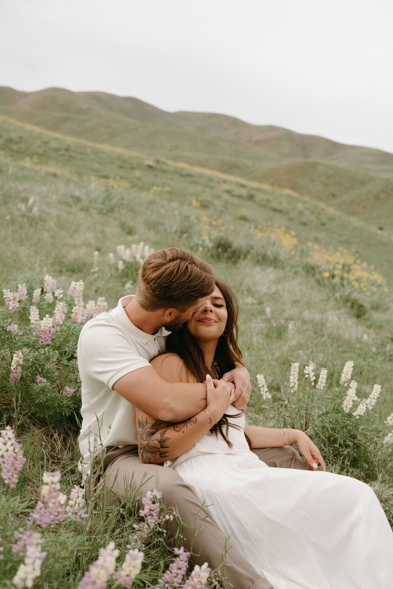 A couple shares an intimate moment among blooming wildflowers on a hillside during a romantic photo session.