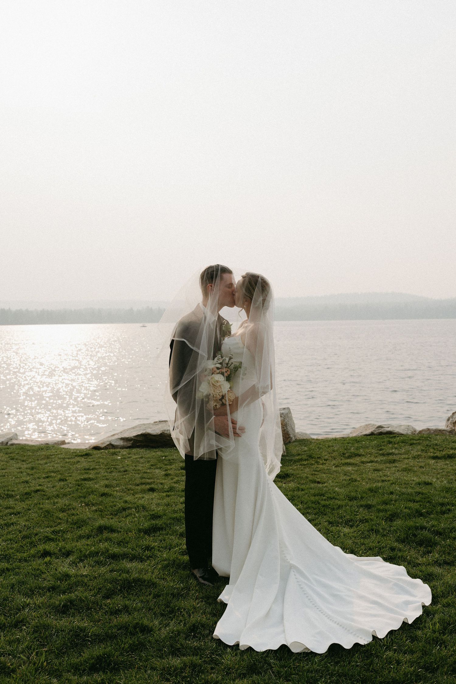A romantic wedding couple shares a tender moment by a lake, with the bride's flowing white dress trailing on green grass.
