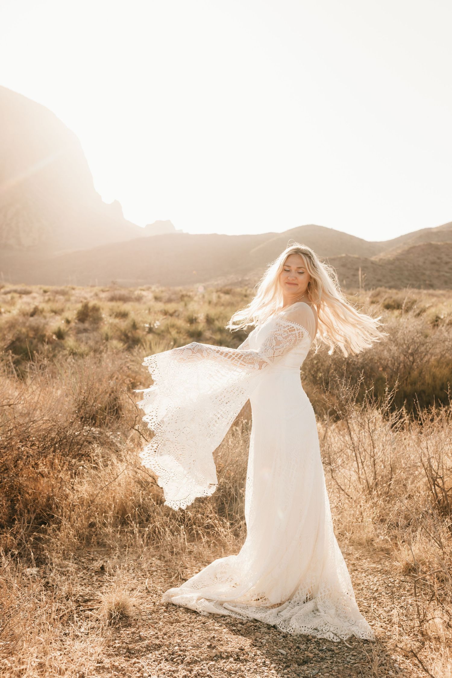 Bride in flowing white dress twirling in desert landscape during sunset.