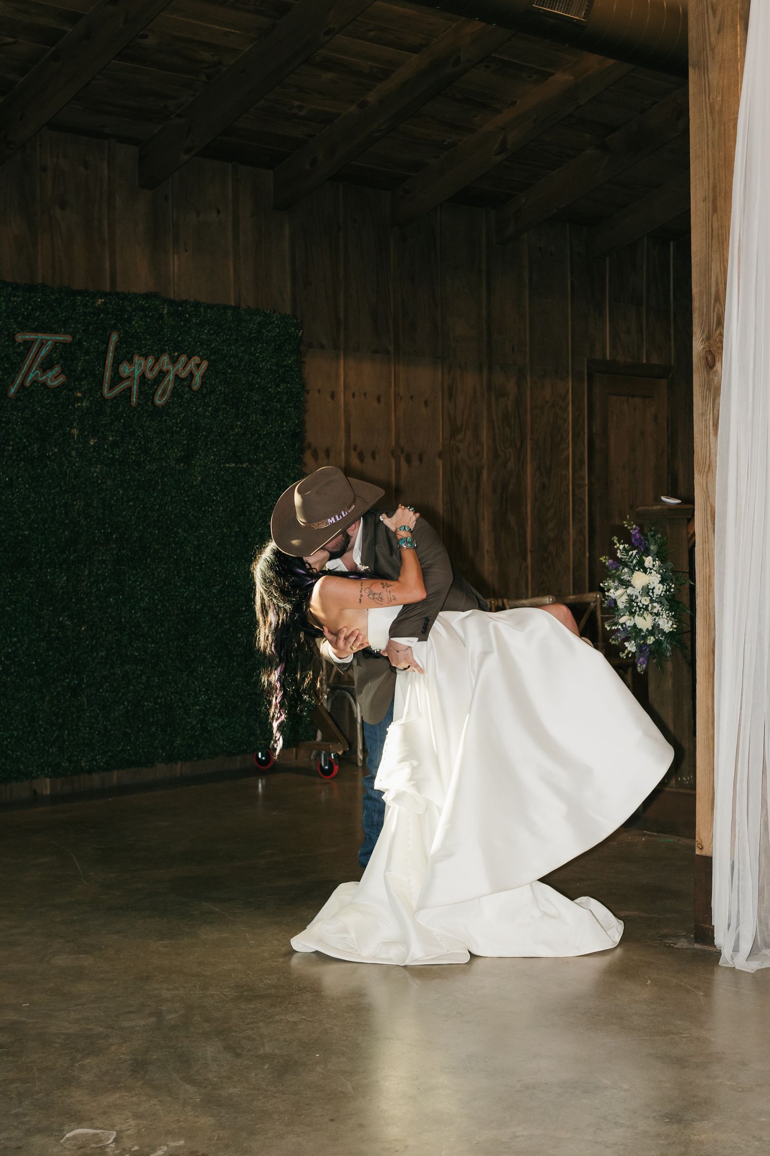 A romantic dip kiss at a rustic barn wedding venue with a green wall backdrop and wooden decor.