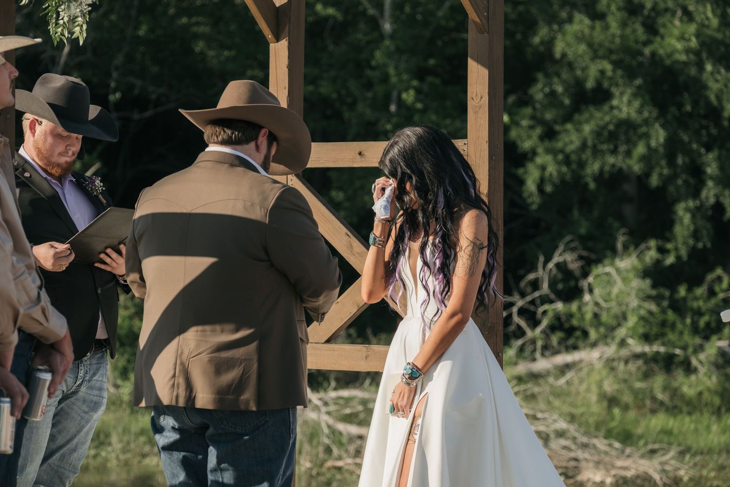 A couple in western attire stands under a wooden pergola during a rustic outdoor wedding ceremony.