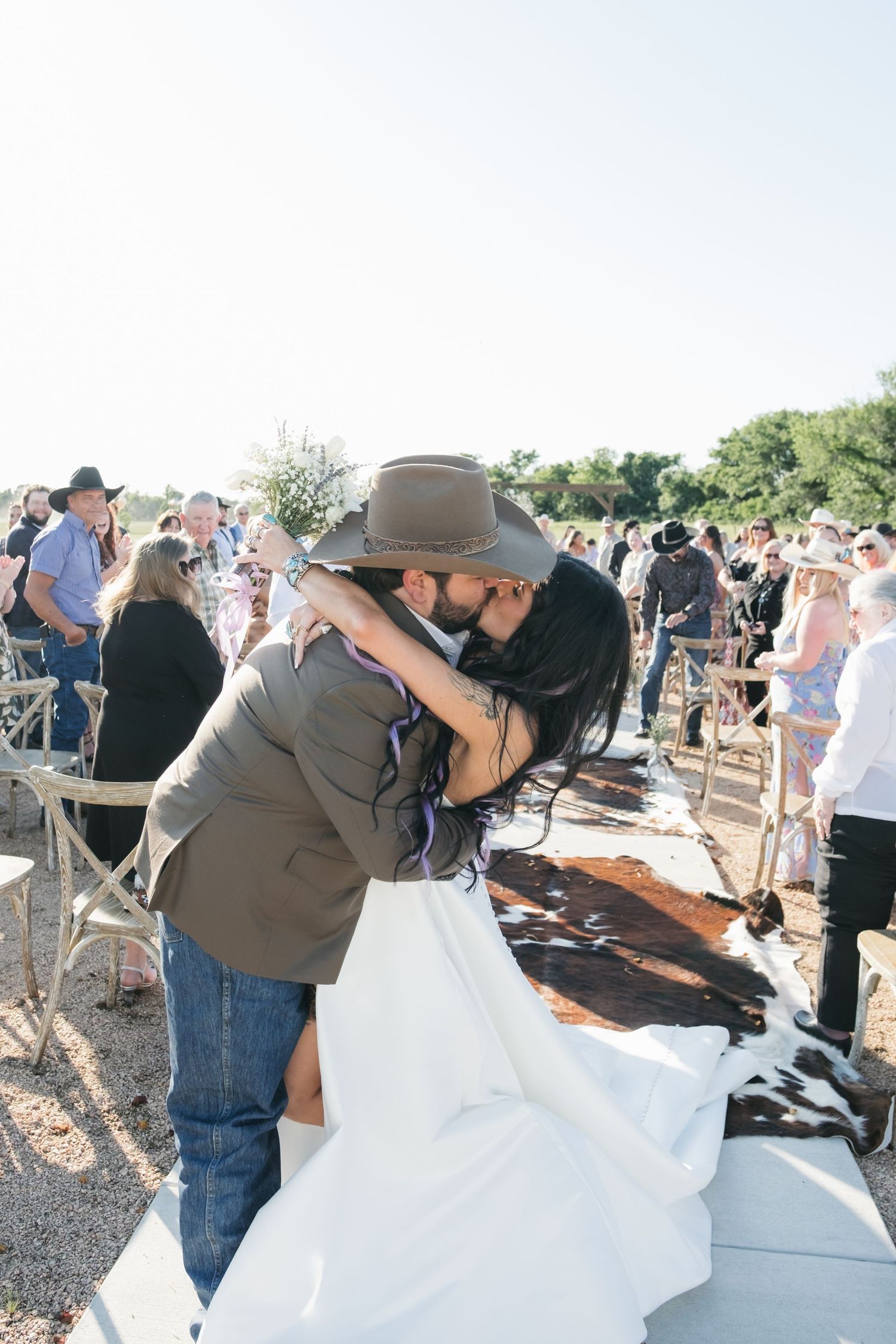 A couple shares their first kiss at a rustic outdoor wedding ceremony surrounded by guests.