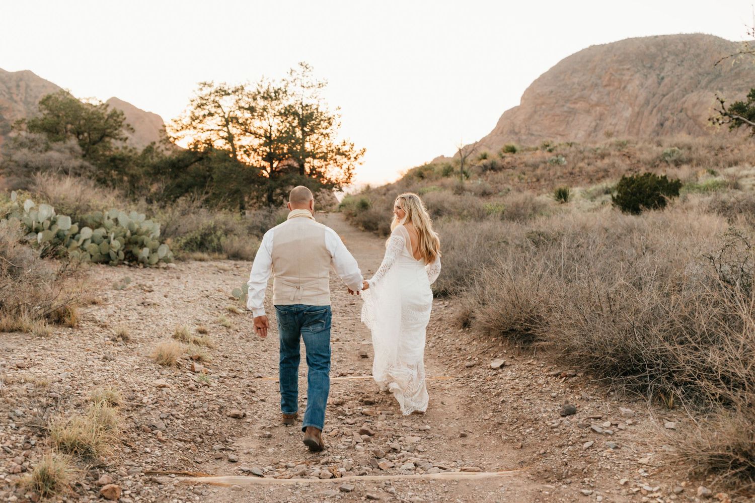 Couple walking hand in hand on desert trail during golden hour with mountains in background.