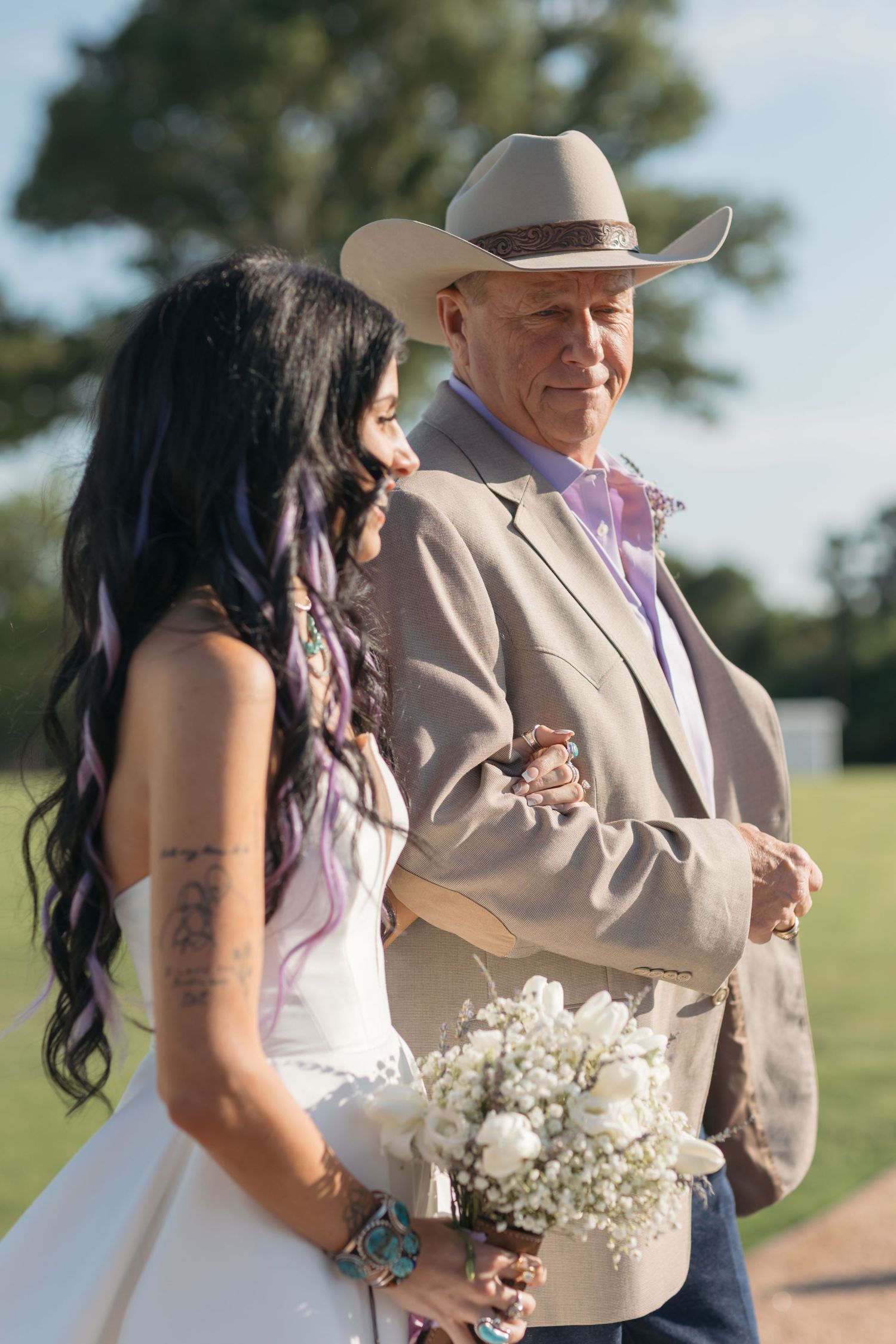A person in a white bridal gown holding baby's breath bouquet stands next to someone wearing a cowboy hat and suit outdoors.