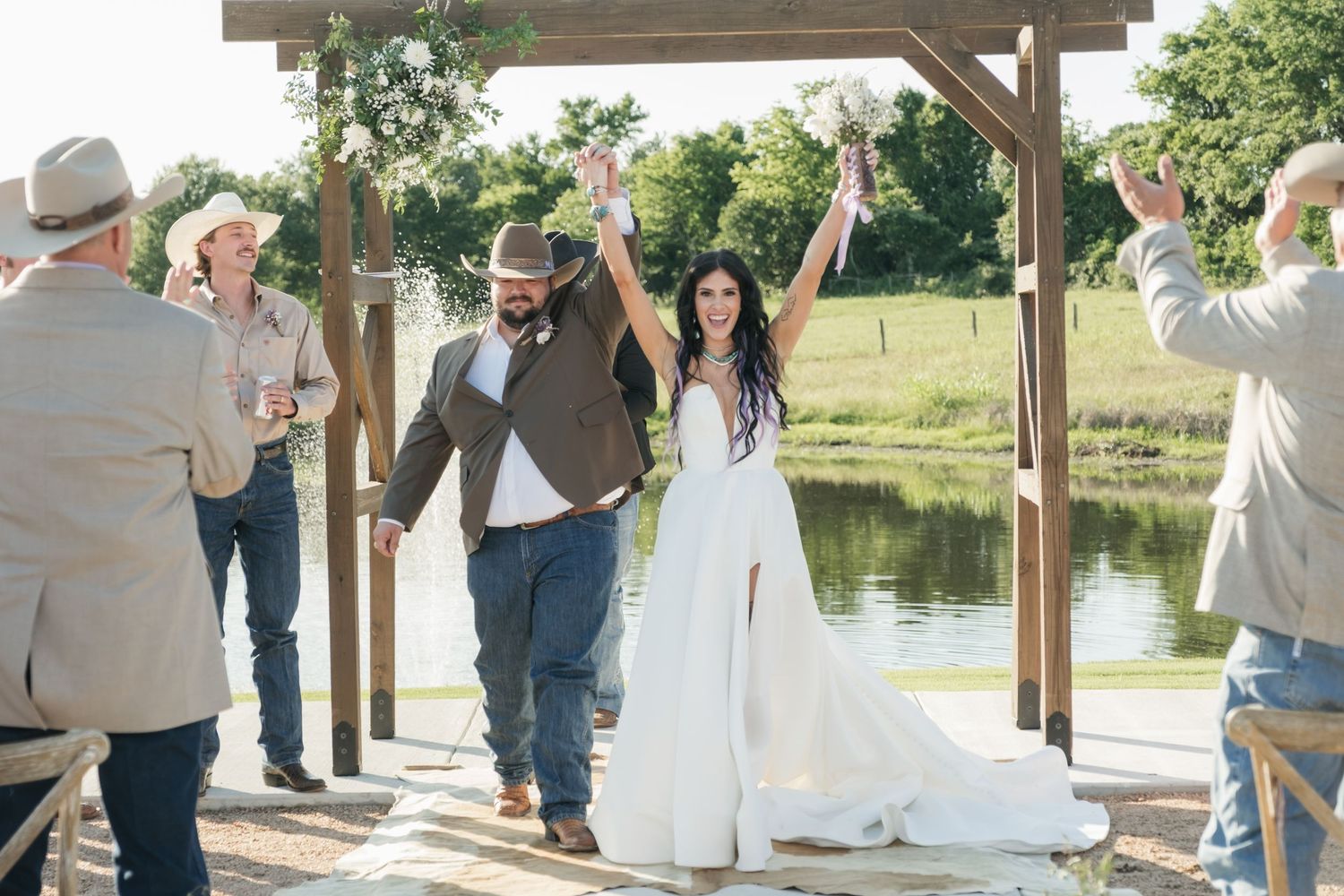 A rustic outdoor wedding ceremony takes place under a wooden pergola next to a tranquil pond in a country setting.