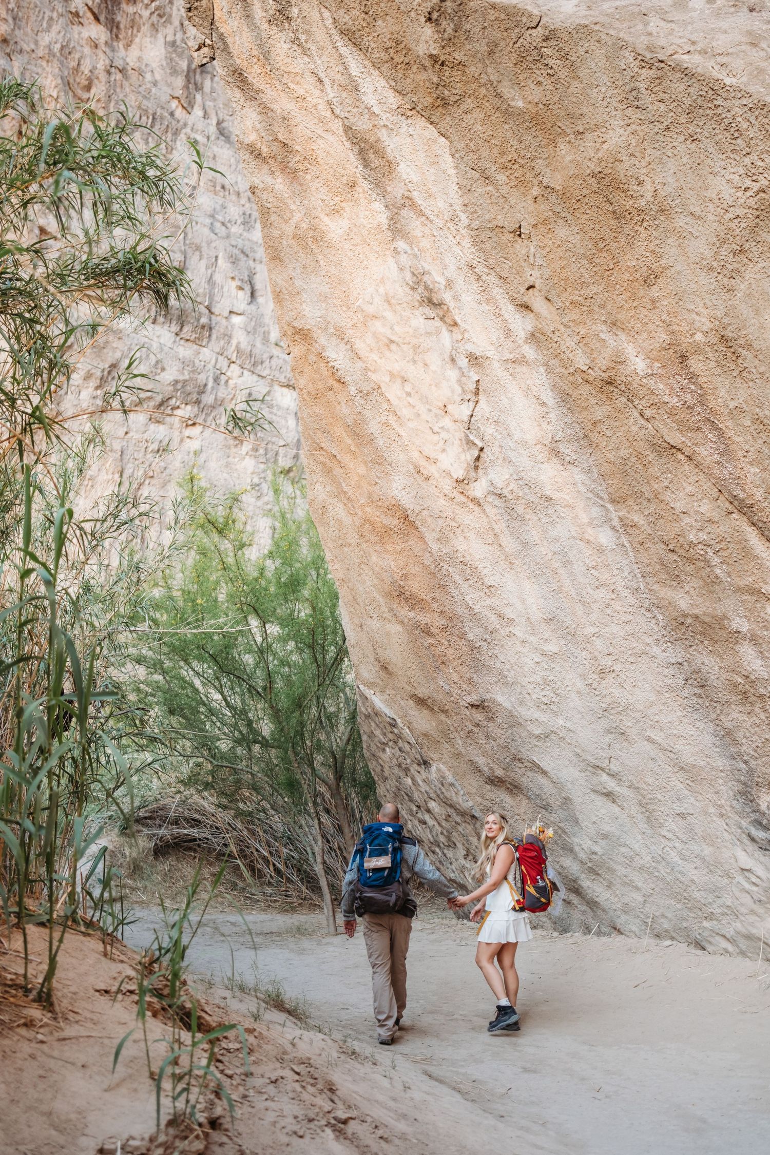 Two hikers walk along a narrow trail between towering sandstone canyon walls.