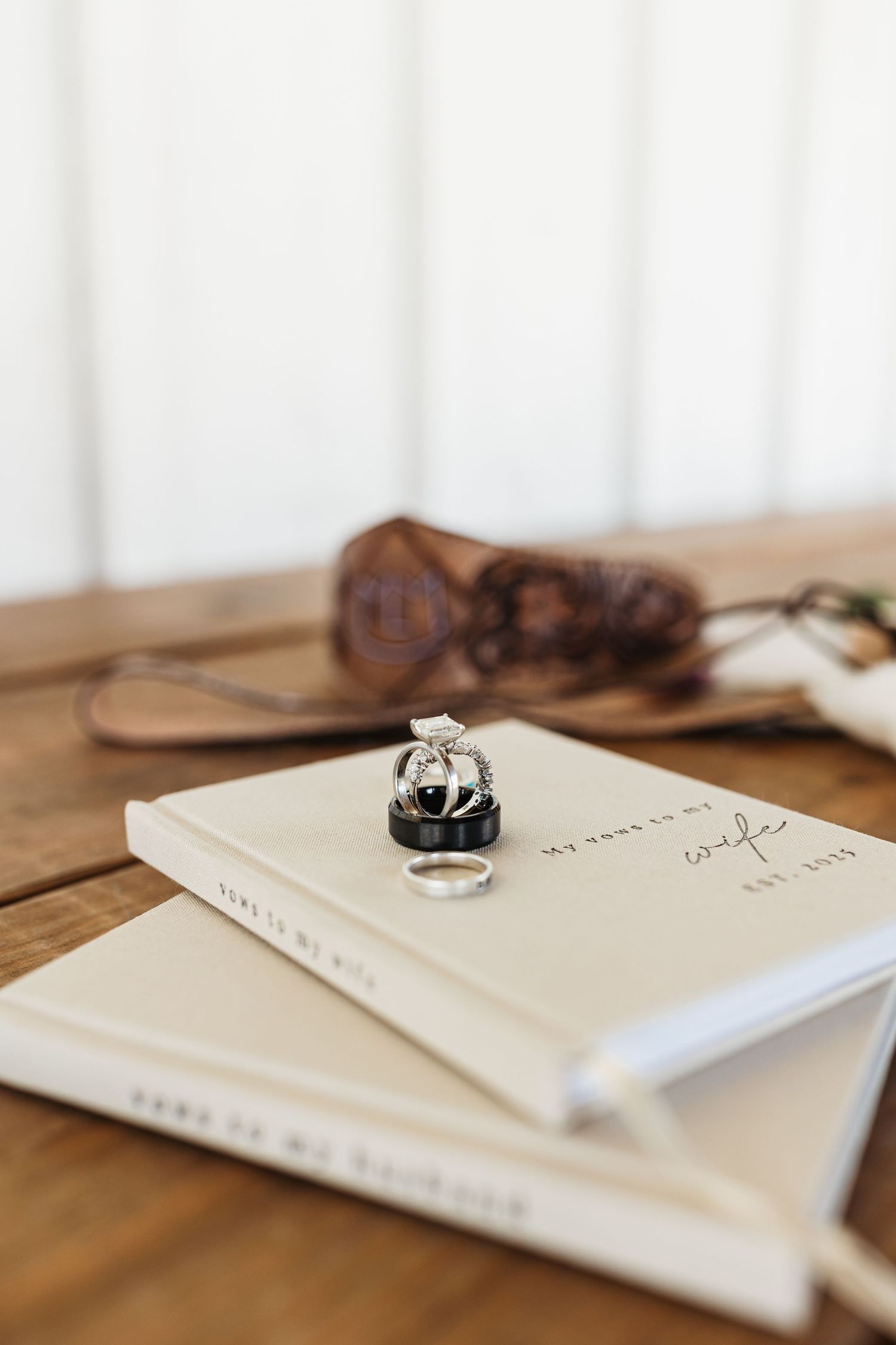 An engagement ring rests on white notebooks with a leather camera strap blurred in the background on a wooden surface.