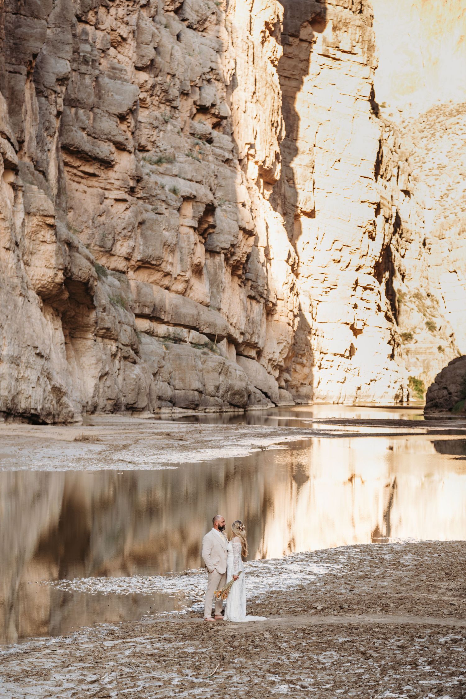 Couple standing by reflective water in dramatic canyon with towering rock walls.