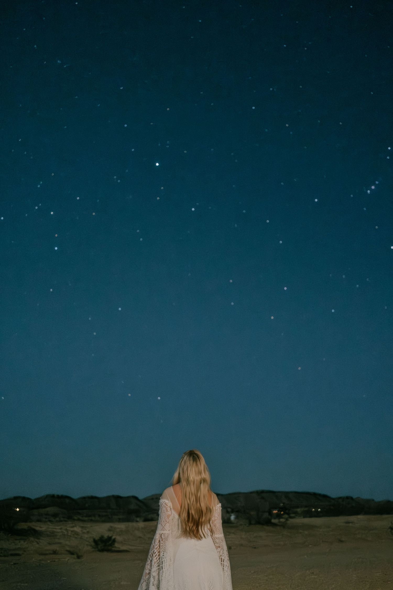 Person in white dress stands beneath starry night sky in desert landscape.