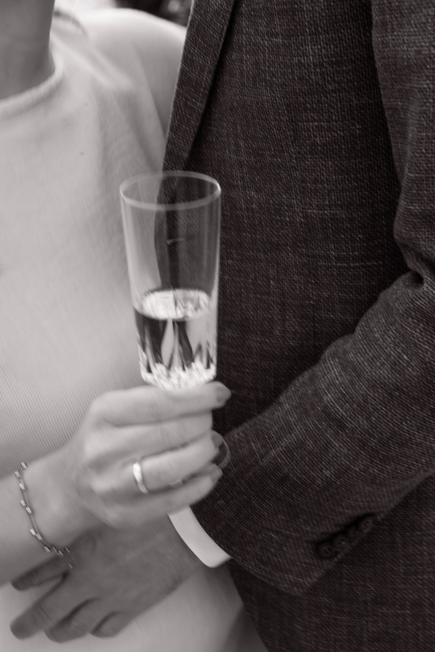 A black and white photo of hands holding a champagne glass, with one hand wearing a ring and bracelet.